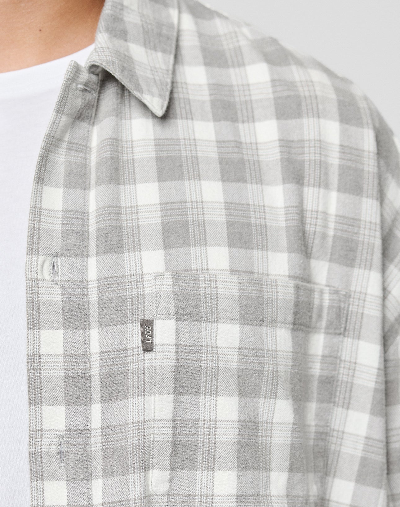 A close-up of a person in the LIVE FAST DIE YOUNG LFDY Light Flannel, a gray and white plaid oversize shirt with a chest pocket and small fabric tag, layered over a white tee; only their chest and neck are visible.