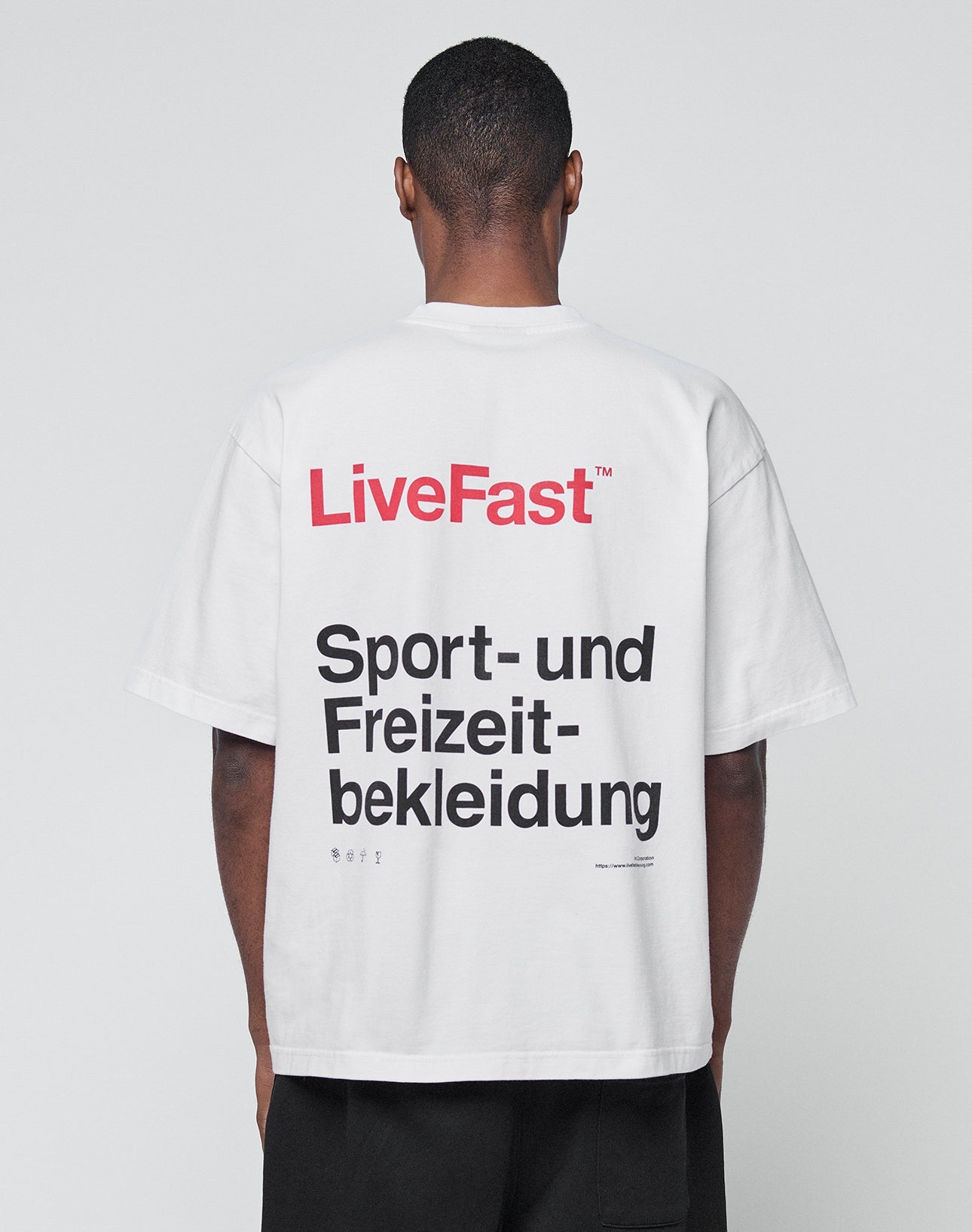A person with short hair is seen from behind in a white Basic Trademark Tee by LIVE FAST DIE YOUNG, made of 100% cotton. The oversize fit features LiveFast in red and bold black Sport- und Freizeitbekleidung on the back.