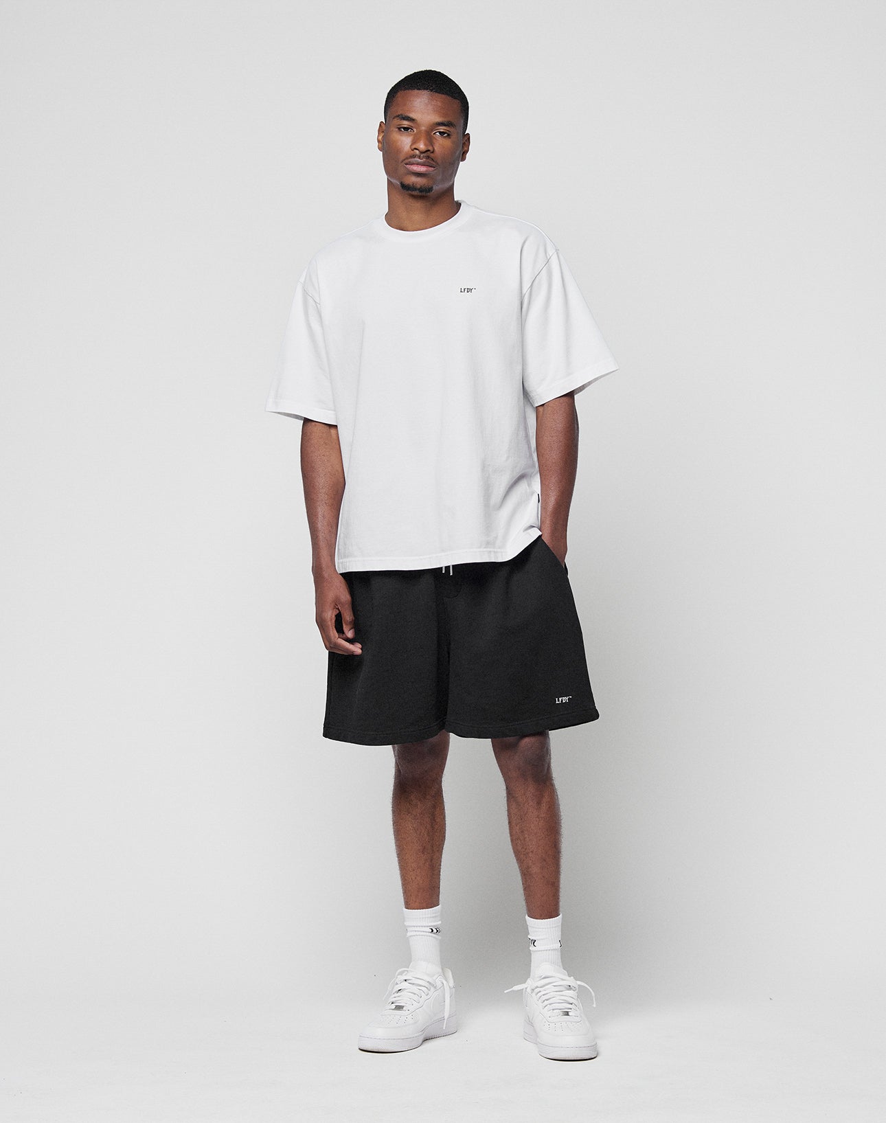 A man stands against a plain white background wearing the LIVE FAST DIE YOUNG Basic Trademark Tee in an oversize fit, paired with black shorts, white socks, and white sneakers. One hand is relaxed in his pocket.