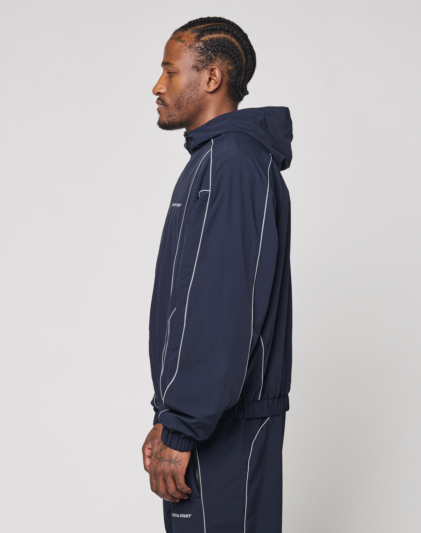 A man with braided hair in profile wears the LIVE FAST DIE YOUNG Live Fast Trackjacket in navy blue, featuring white piping, reflective print, elastic cuffs, and zip pockets.