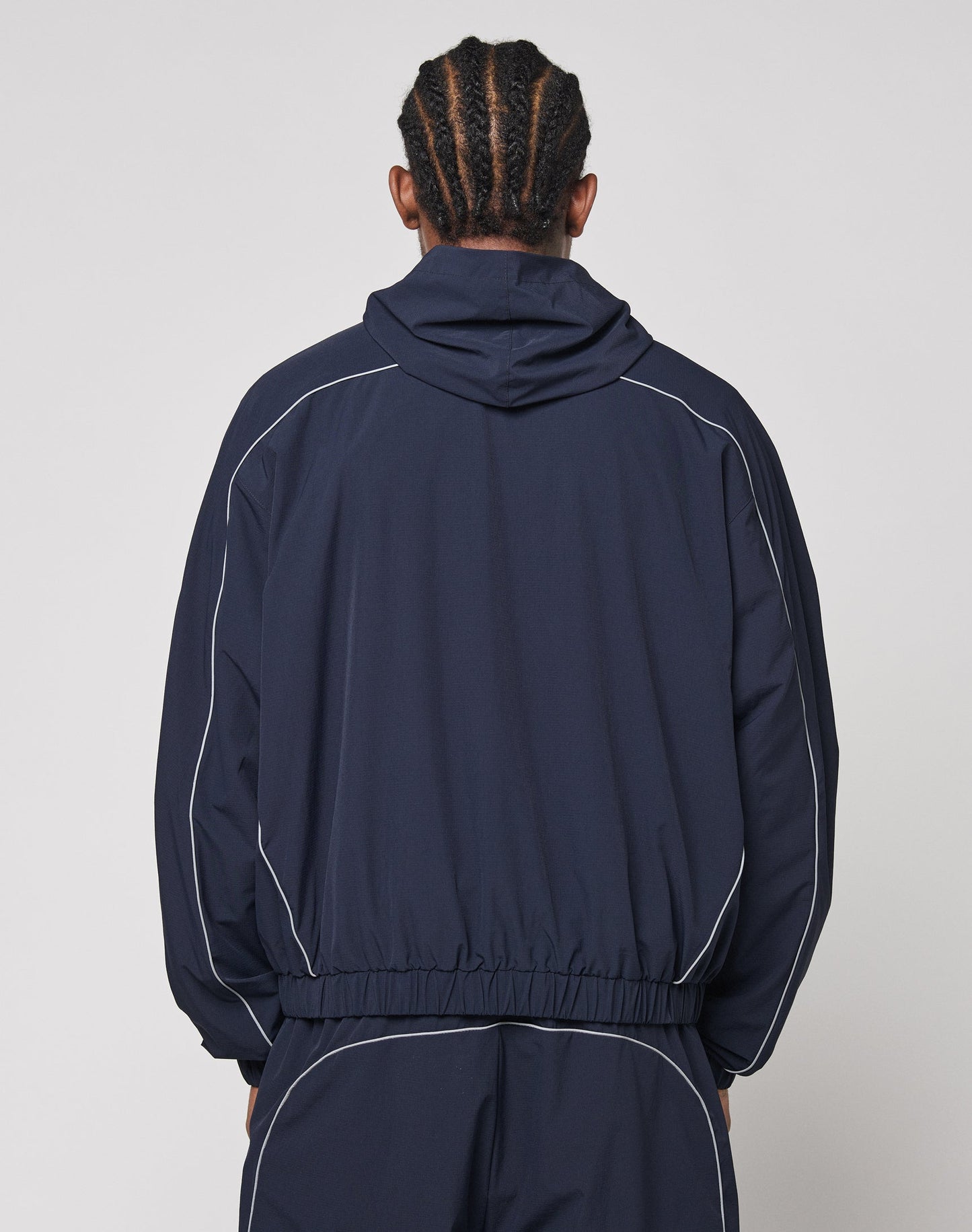 A person with braided hair, seen from the back, wears the LIVE FAST DIE YOUNG Live Fast Trackjacket with white piping, elastic cuffs, zip pockets, and matching pants against a simple light background.