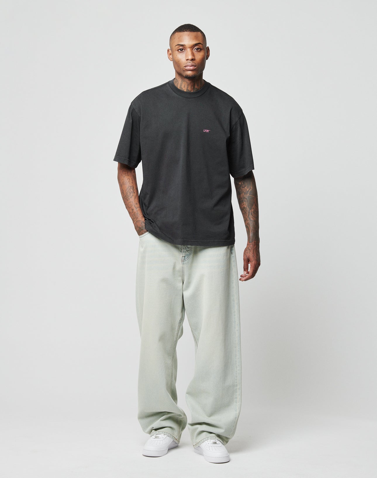 A man stands facing forward against a plain background, wearing a loose black Baggy Fit T-shirt, LIVE FAST DIE YOUNG LFDY Baggy Denim jeans in light gray with a subtle Flag-Label, and white sneakers. His arms and neck are tattooed.