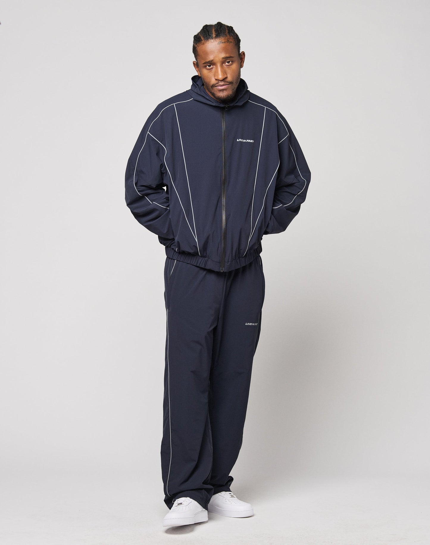 A man wears the LIVE FAST DIE YOUNG Live Fast Trackjacket, featuring navy blue fabric with white reflective print, elastic cuffs, and zippered pockets. He pairs it with white sneakers and stands against a plain background with hands in his pockets.