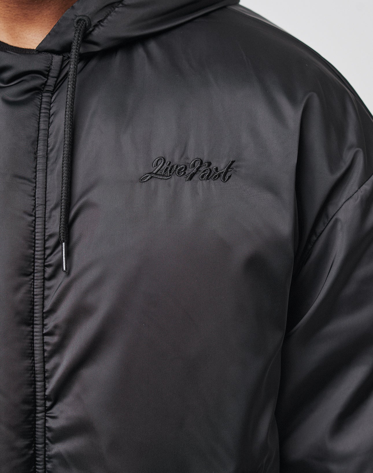 Close-up of the LFDY Winter Bomber Jacket by LIVE FAST DIE YOUNG, featuring a black hood with drawstrings and Live Fast embroidered in black script on the chest. Only part of the model in this Oversize Fit style is visible.