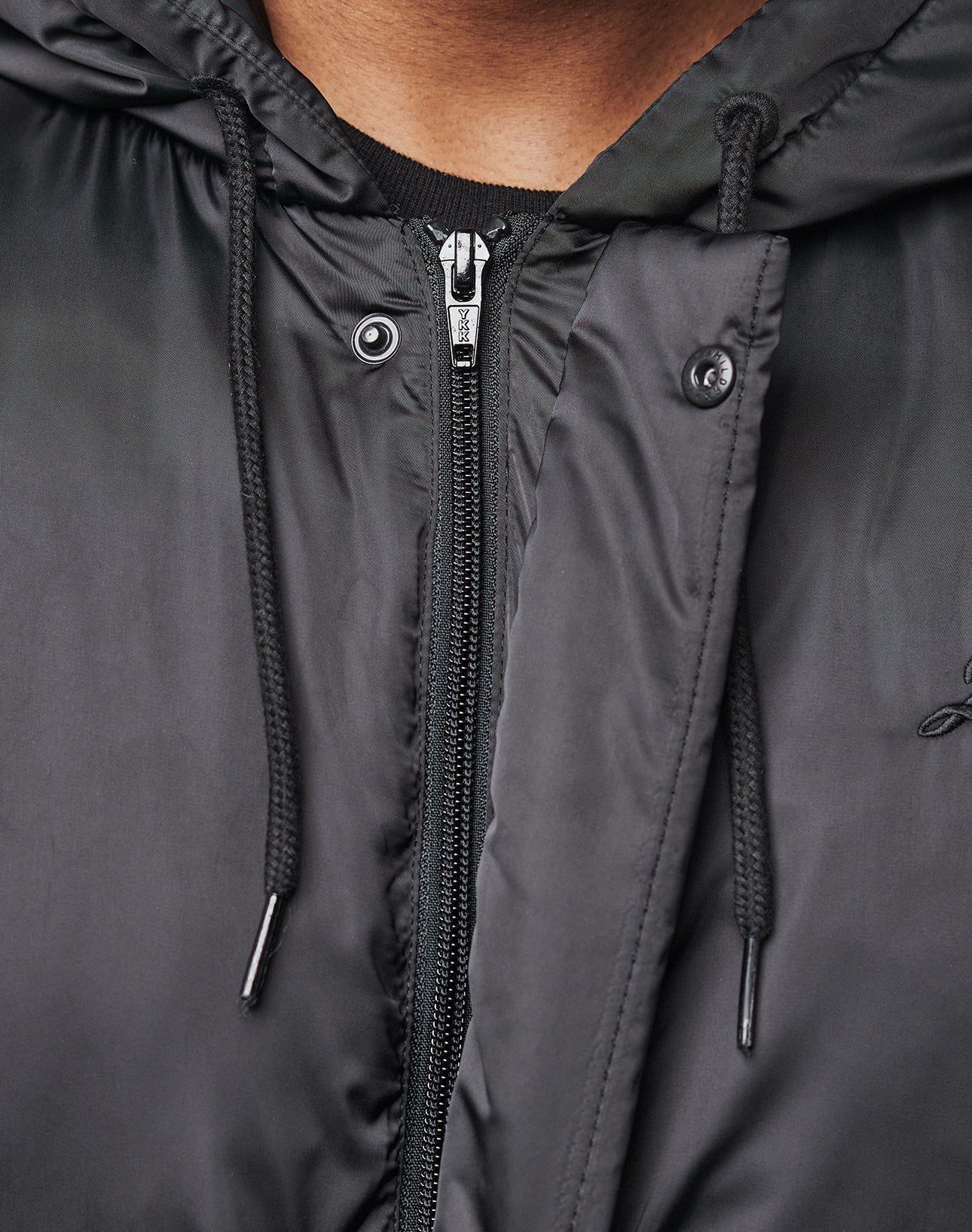 Close-up of a person in the LIVE FAST DIE YOUNG LFDY Winter Bomber Jacket, showing only the lower face, neck, and chest. The shiny black polyester features a hood with metal eyelets and toggles for an Oversize Fit look.