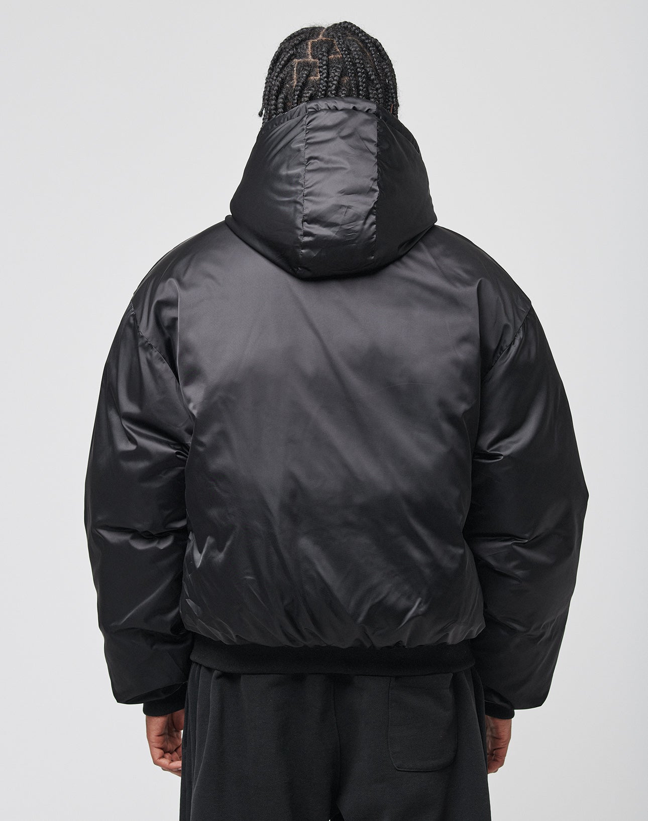 A person with braided hair, seen from the back against a plain light gray background, is wearing an oversize fit shiny black LFDY Winter Bomber Jacket by LIVE FAST DIE YOUNG with a hood.