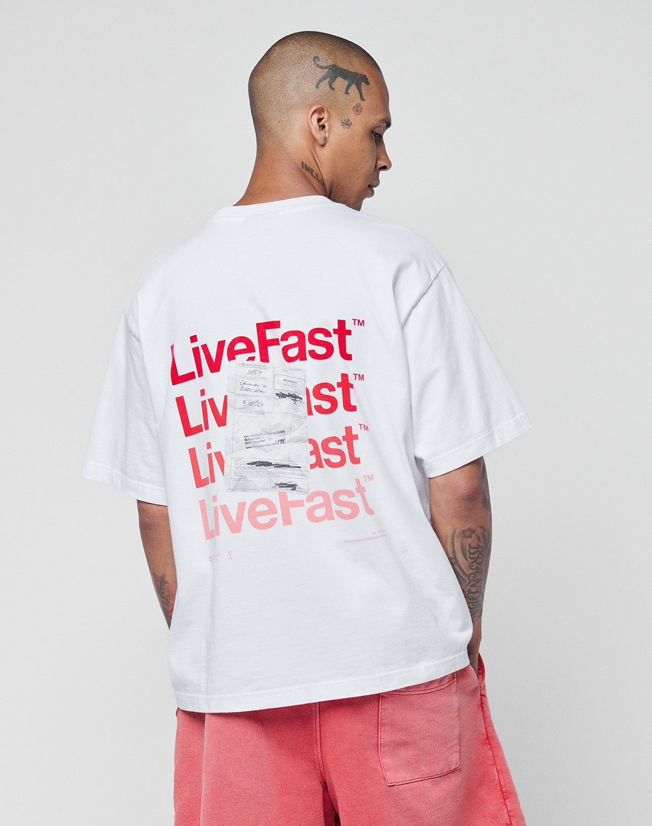 A tattooed, shaved-head person wears the LIVE FAST DIE YOUNG Basic Evidence Tee—an oversized white 100% Baumwolle T-shirt with a bold red LiveFast backprint—paired with pink shorts, standing against a plain gray background.