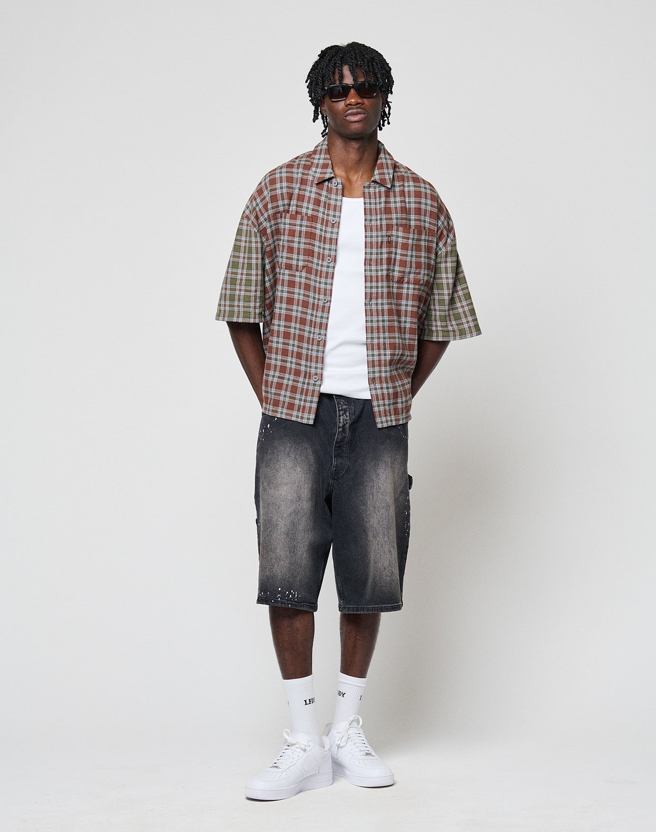 A man with short dreadlocks and sunglasses wears the LIVE FAST DIE YOUNG LFDY Light Flannel shirt over a white tee, paired with knee-length faded black denim shorts, white socks, and sneakers, standing against a plain light background.