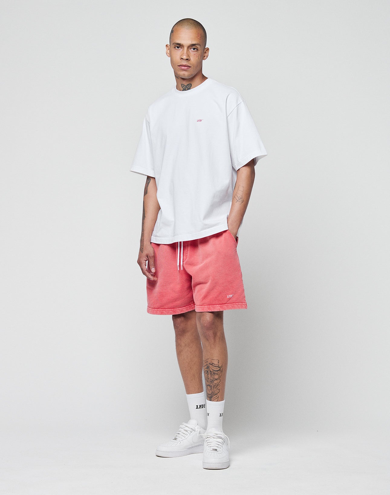 A person stands against a plain backdrop wearing the LIVE FAST DIE YOUNG Basic Evidence Tee—an oversize, loose white shirt made of 100% Baumwolle—paired with pink drawstring shorts, white crew socks, sneakers, and showing tattoos on forearm and shin.