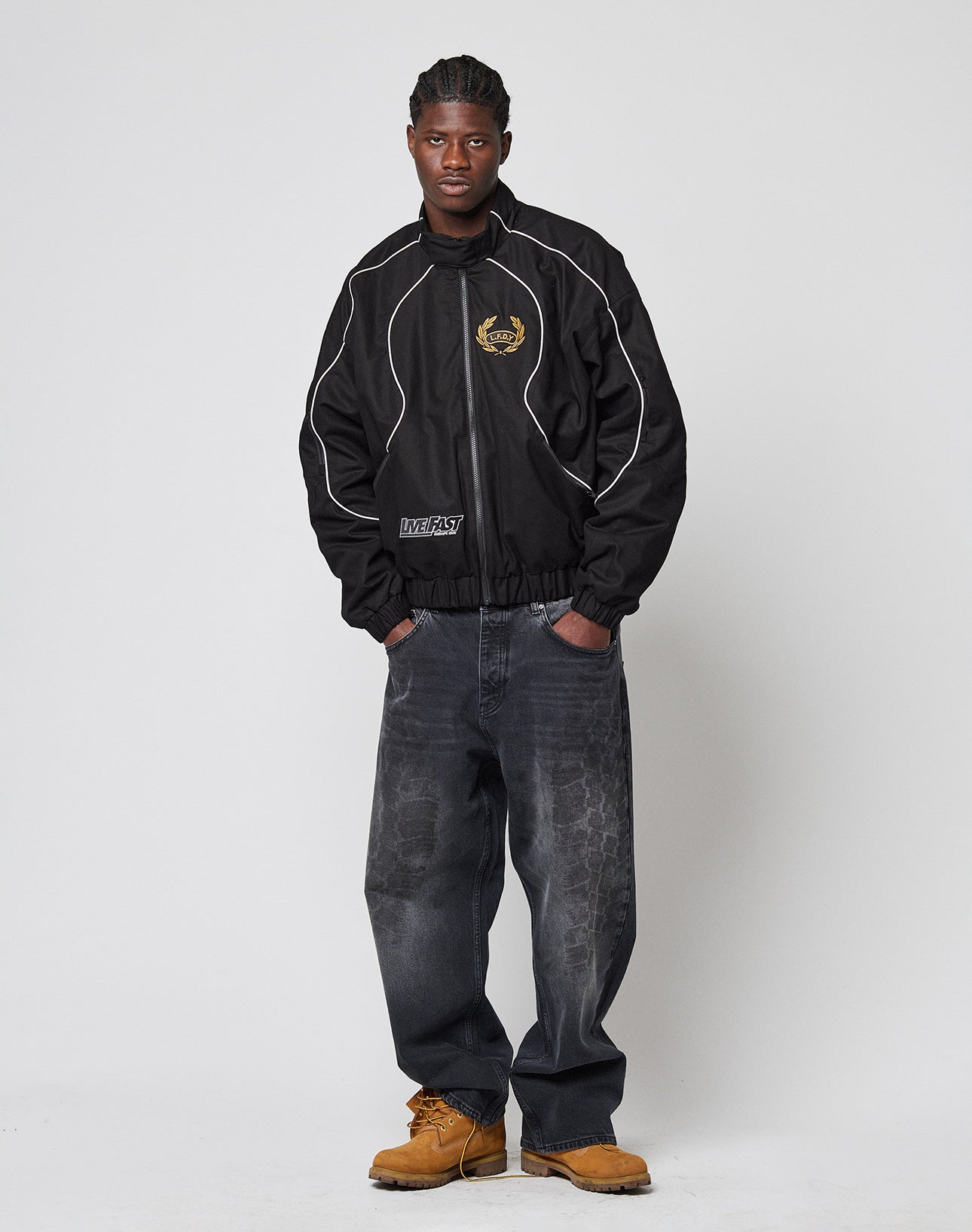 A man stands against a plain white background wearing the LFDY Canvas Jacket by LIVE FAST DIE YOUNG, paired with faded baggy jeans and tan work boots. He has short braided hair and his hands are in his pockets.