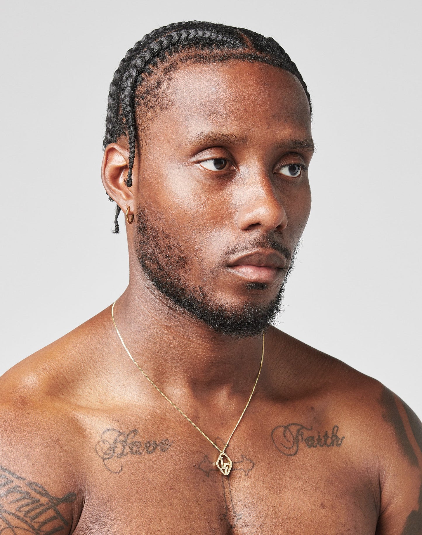 A man with braided hair and a short beard looks slightly to the side, wearing the LF Chain by LIVE FAST DIE YOUNG and a small hoop earring. Have Faith is tattooed on his chest. The background is plain light gray.