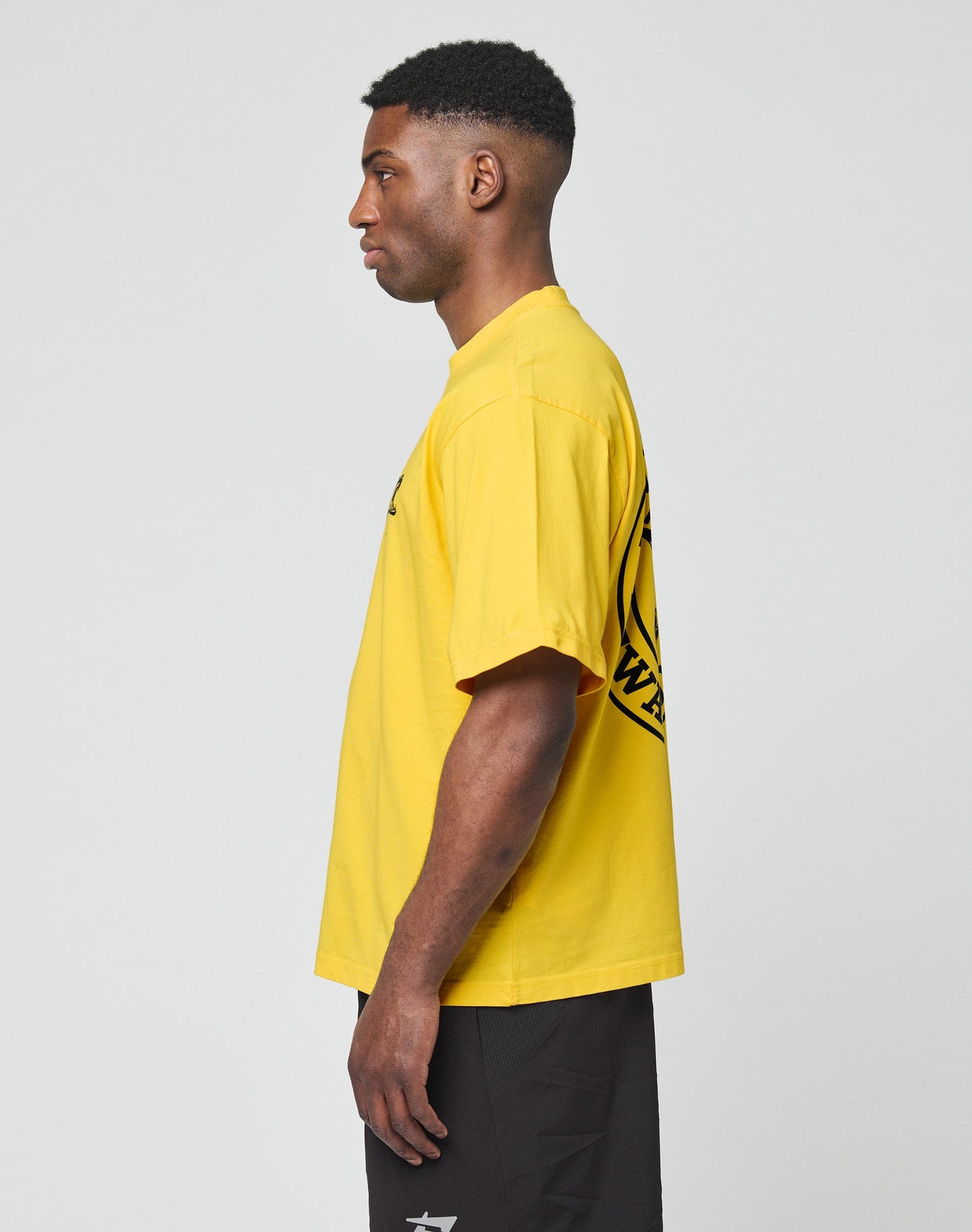 A person in profile wears the LIVE FAST DIE YOUNG LFDY Circle Dog Tee, a bright yellow 100% Baumwolle t-shirt with a back graphic, and black shorts, standing against a plain light gray background.