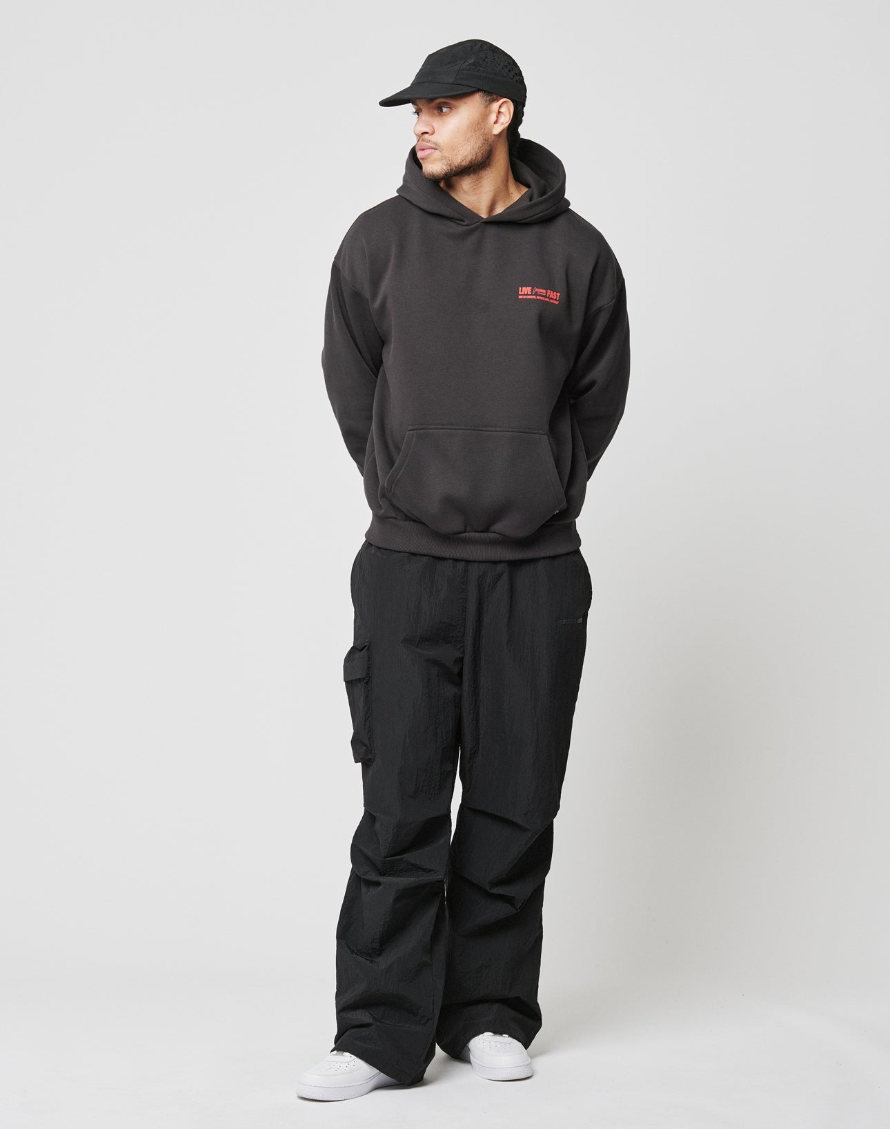 A man models the LIVE FAST DIE YOUNG Hit and Run Hooded, an oversized black hoodie with red text, paired with black cargo pants, a black cap, and white sneakers against a plain white background. He looks left with his hands in his pockets.