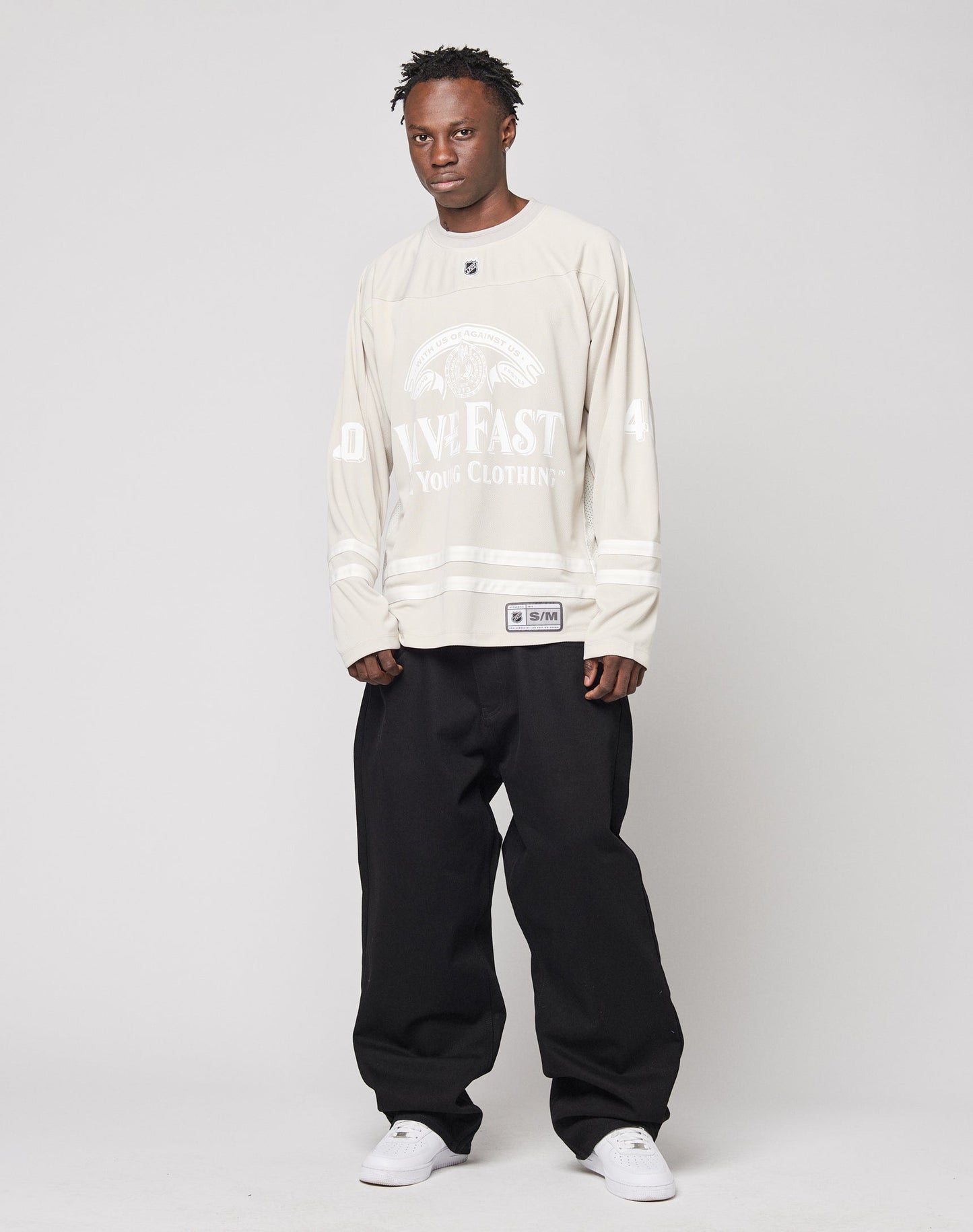 A man stands against a plain background wearing the LFDY Hockey Jersey by LIVE FAST DIE YOUNG with beige long sleeves, white lettering, and Ärmelprint, paired with baggy black Oversize Fit pants and white sneakers. He looks at the camera neutrally.