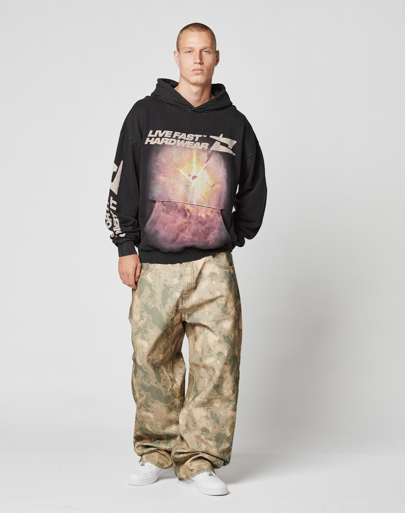 A man stands against a plain background wearing the LIVE FAST DIE YOUNG LFDY Angel Hooded, baggy camo-print pants, and white sneakers, gazing directly at the camera with a neutral expression.
