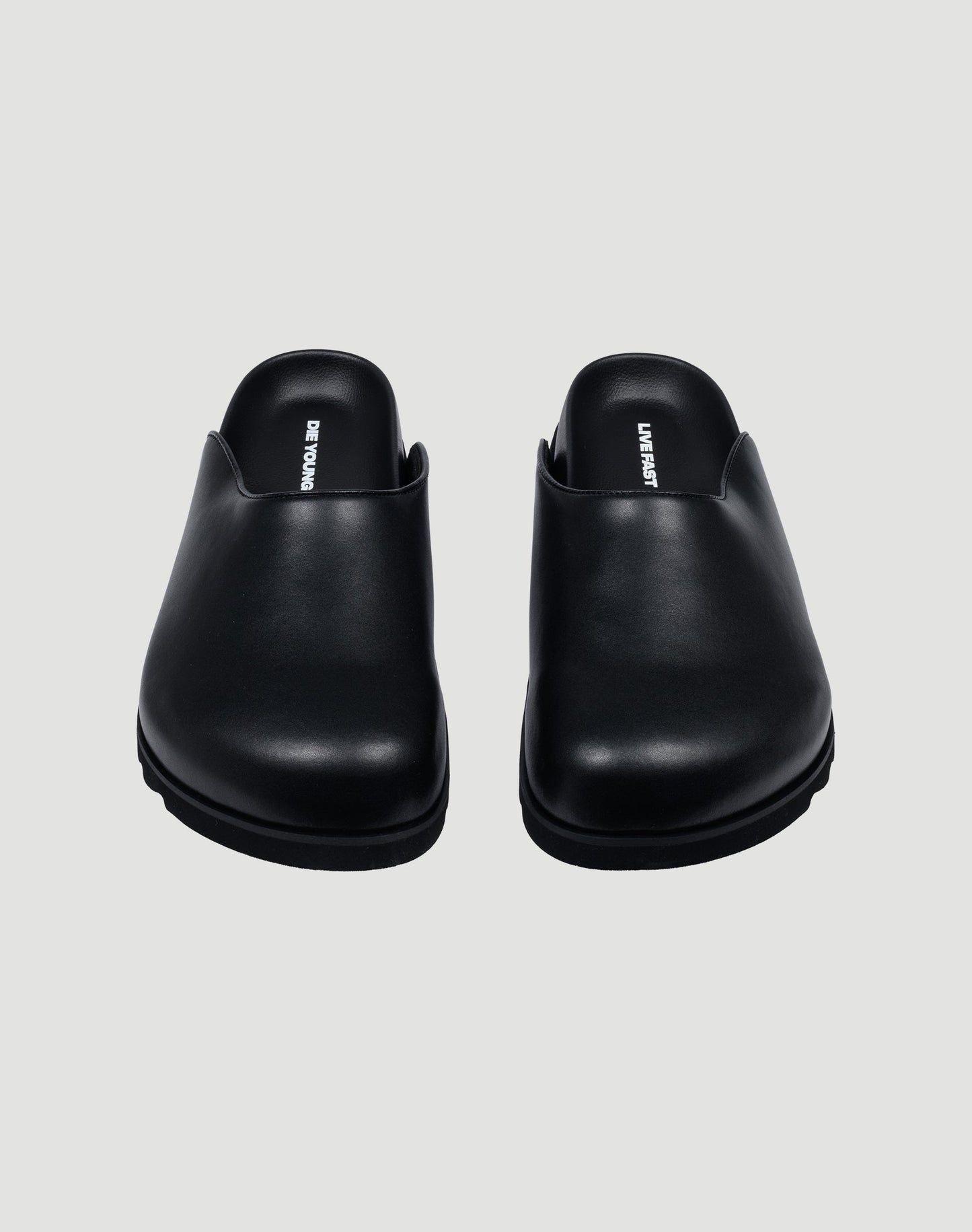 The LFDY Mules by LIVE FAST DIE YOUNG are black slip-on leather shoes with closed rounded toes, schwarze Innenmaterial, and a durable PU sole, shown from the front on a light gray background.