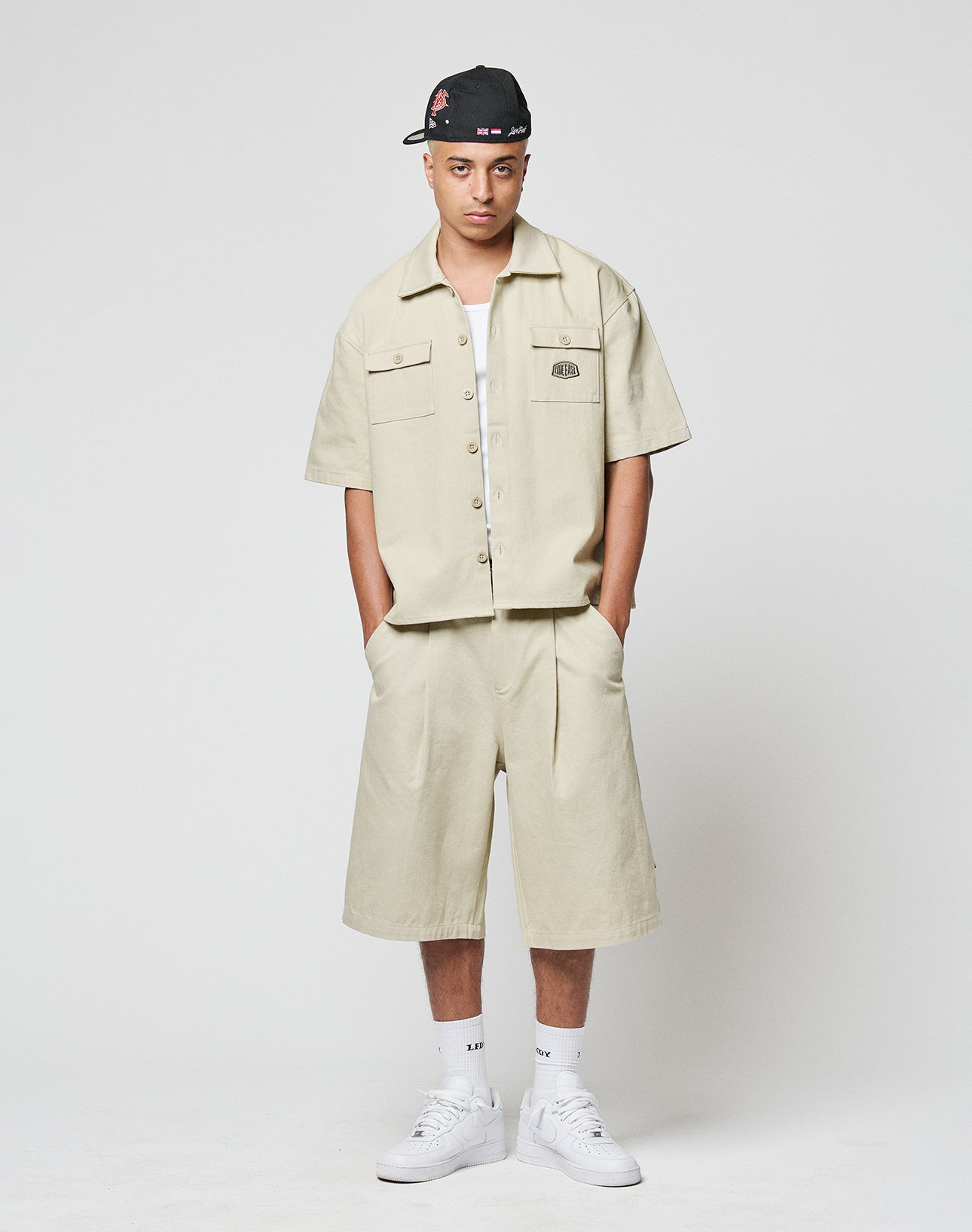 A person stands against a plain background wearing LIVE FAST DIE YOUNGs beige LFDY Twill Shorts, an oversized matching short-sleeve button-up shirt, white socks, white sneakers, and a black cap with red and white designs.