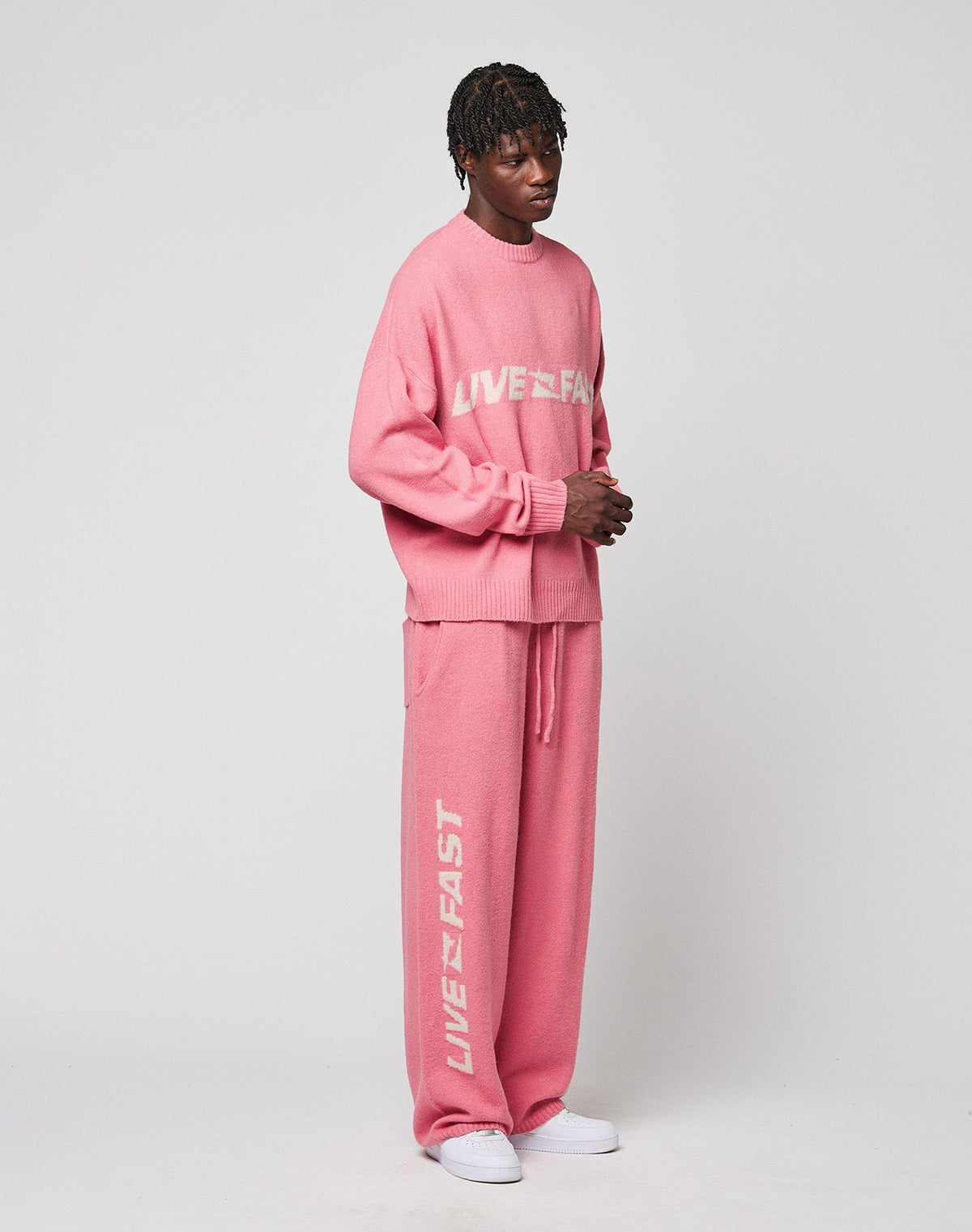 A person poses against a plain background in the LIVE FAST DIE YOUNG Live Fast Boucle Pants with "LIVE FAST" in white, featuring an elastic waistband and straight leg, styled with a matching pink sweater and white sneakers.