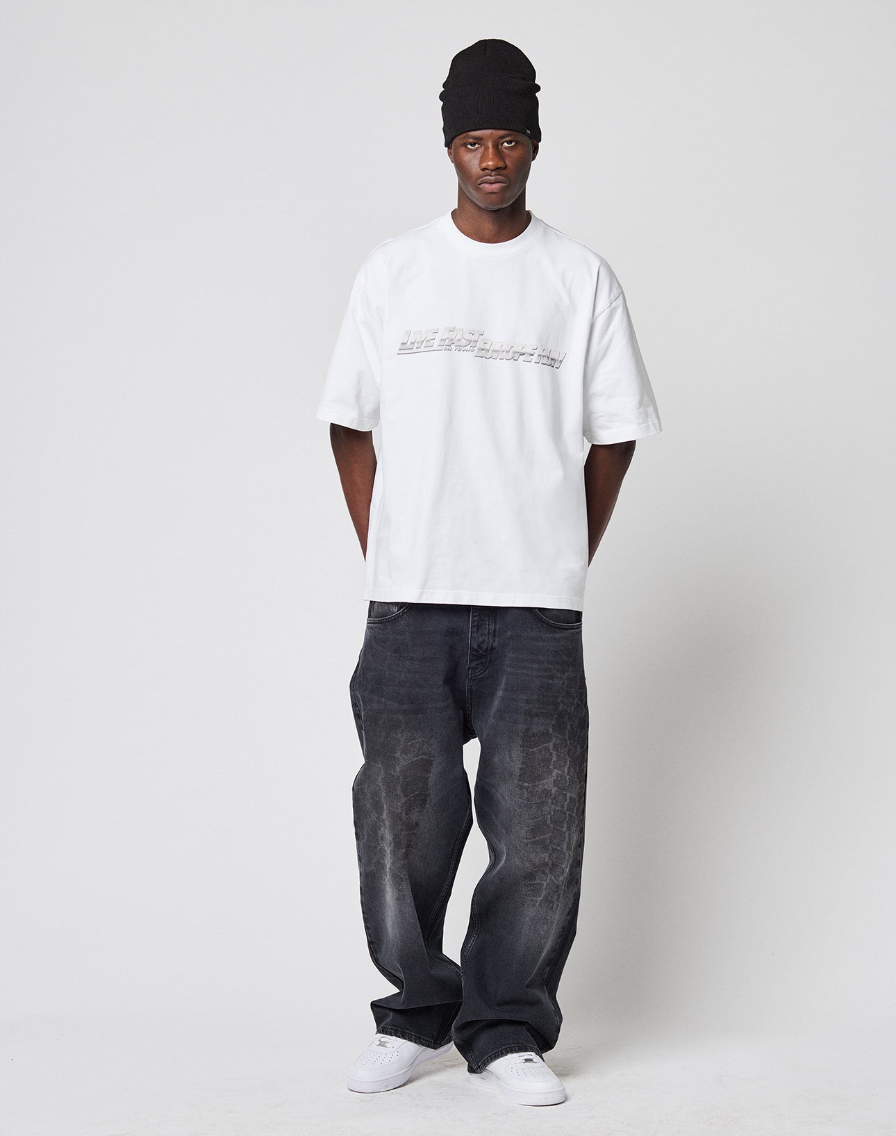 A person stands against a plain background, wearing a black beanie, loose dark jeans, white sneakers, and a LIVE FAST DIE YOUNG LFDY Europe Run Tee—an oversized, white 100% Baumwolle T-shirt with text—hands behind their back and a neutral expression.