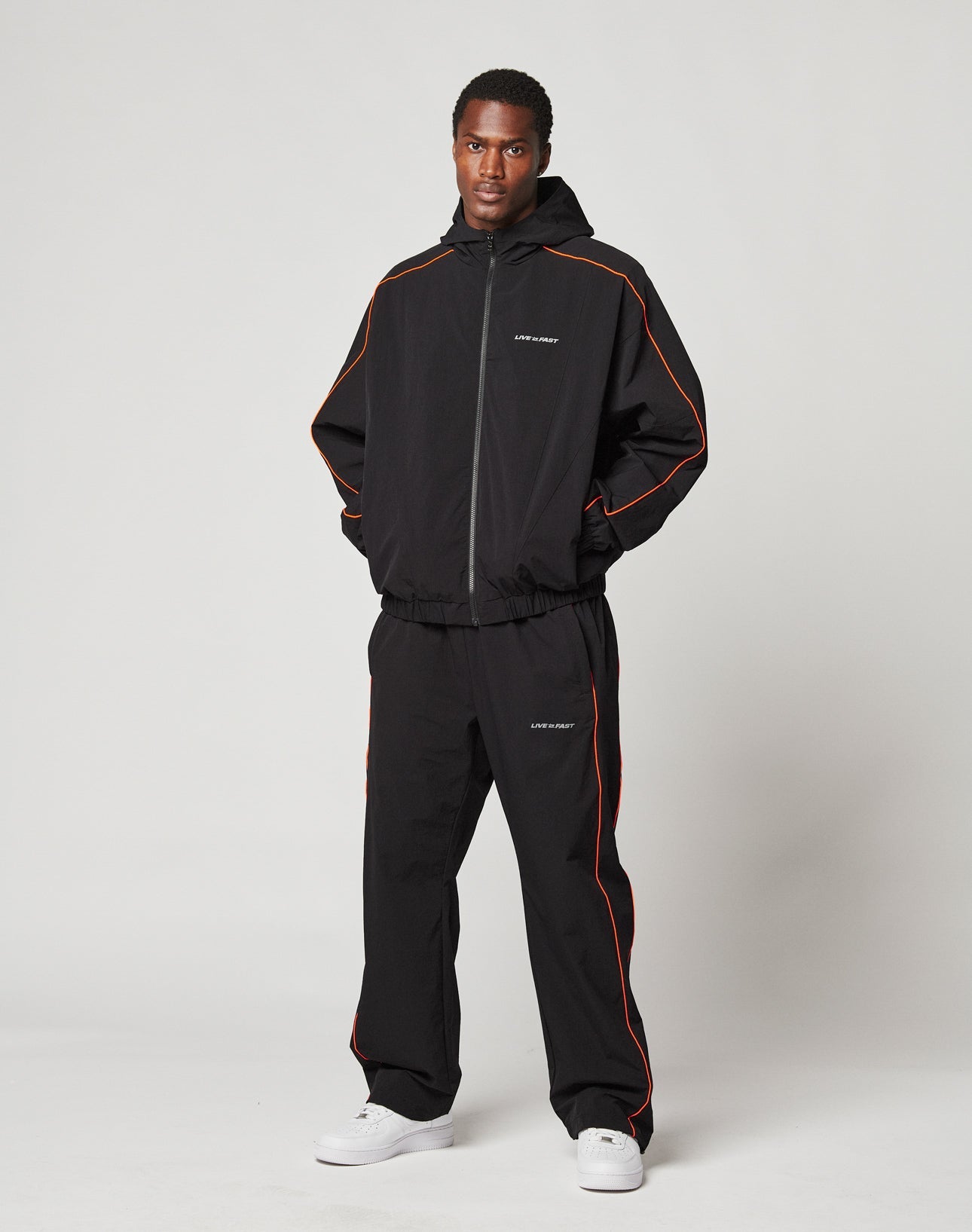 A person stands facing forward in the LIVE FAST DIE YOUNG Live Fast Trackjacket, black with orange piping, reflektierender Druck, elastische Bündchen, hood, white text on chest and leg, hands in pockets, white sneakers, plain light background.