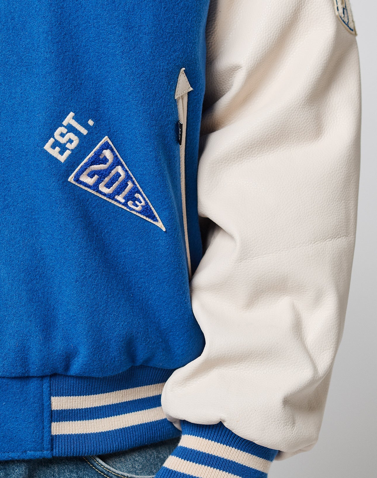 Close-up of the LIVE FAST DIE YOUNG LFDY x Schott NYC Varsity Jacket, showing EST. 2013 white and blue chenille patches, blue and cream striped ribbing, a zippered pocket detail, and cowhide leather sleeves for a premium look.
