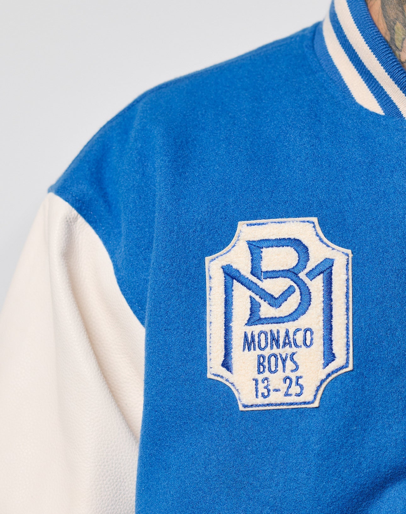 Close-up of the LIVE FAST DIE YOUNG x Schott NYC Varsity Jacket in blue and white, featuring cowhide leather sleeves and a chenille Monaco Boys 13-25 patch with a stylized MB logo on the chest.