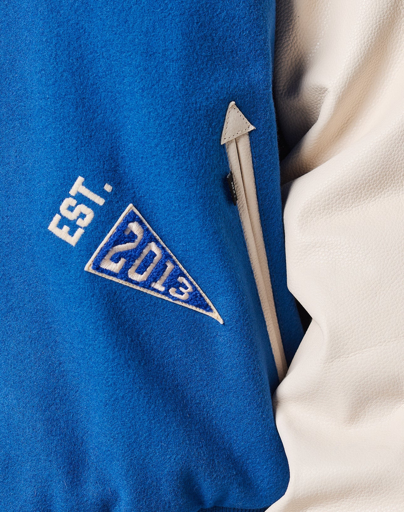 Close-up of the LFDY x Schott NYC Varsity Jacket by LIVE FAST DIE YOUNG, showcasing cowhide leather sleeves, a zippered pocket, and an embroidered blue triangle patch with white EST. 2013 text.