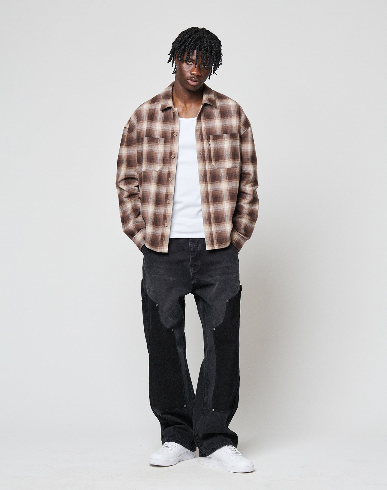 A man with short dreadlocks stands against a plain background wearing a LIVE FAST DIE YOUNG Vintage Flannel over a white T-shirt, paired with loose black pants and white sneakers. His hands are in his pockets.
