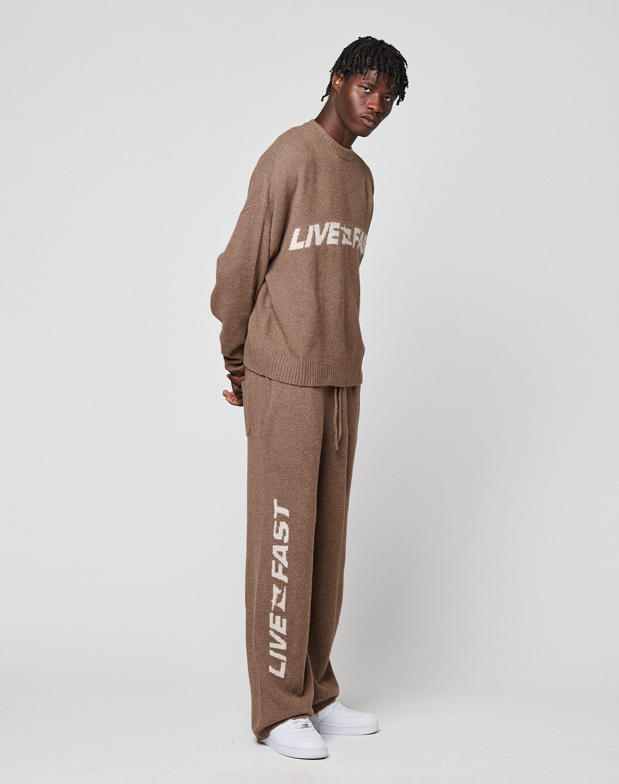A man wears LIVE FAST DIE YOUNG's Live Fast Boucle Pants with a matching brown sweater, both featuring white "LIVE FAST" print. The outfit has gerade Bein pants with an Elastikbund, styled simply with white sneakers against a white backdrop.