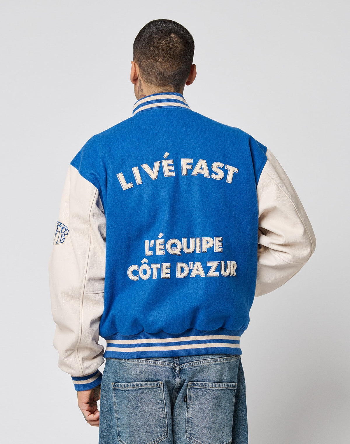 Someone stands facing away from the camera in a blue and white LFDY x Schott NYC Varsity Jacket by LIVE FAST DIE YOUNG, featuring cowhide leather sleeves and LIVE FAST LÉQUIPE CÔTE DAZUR embroidered on the back.