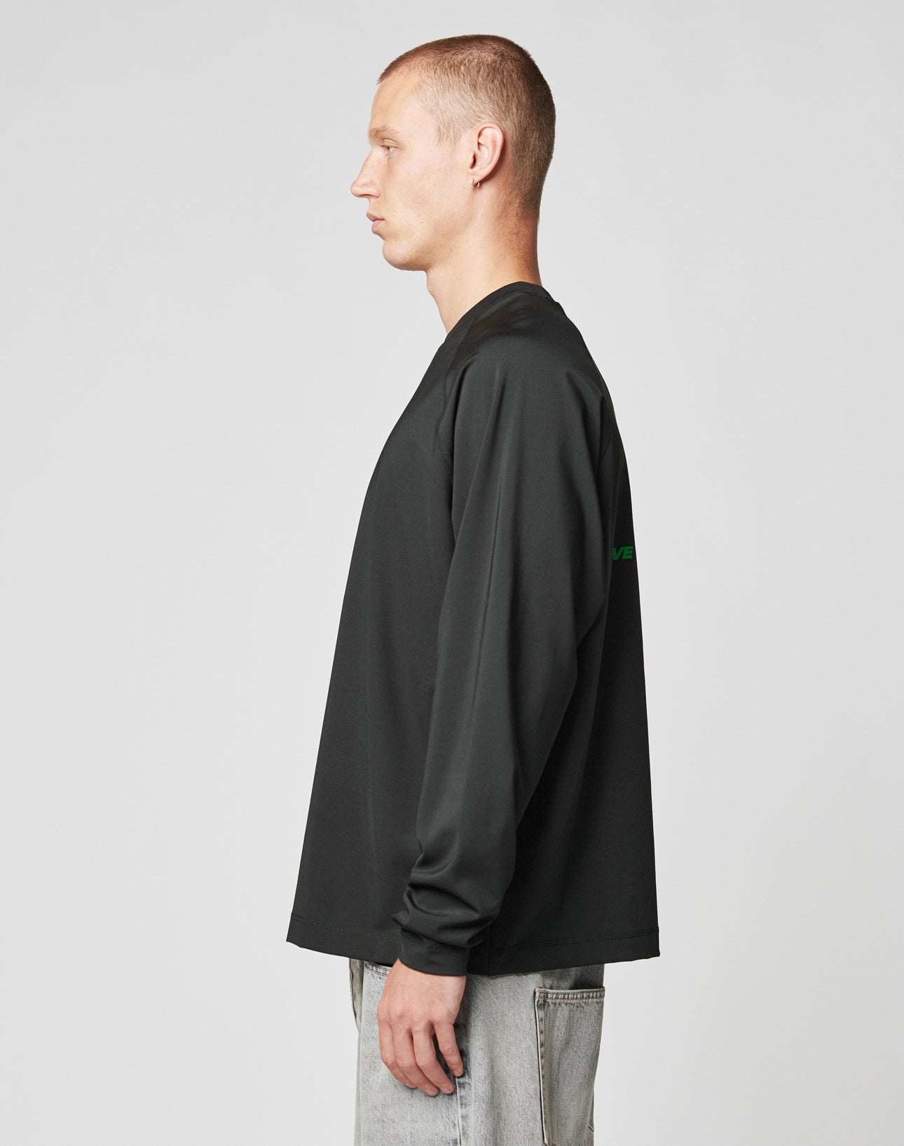A man with short hair stands in profile facing left, wearing a black LFDY Sports Longsleeve by LIVE FAST DIE YOUNG and light gray jeans against a plain light background.