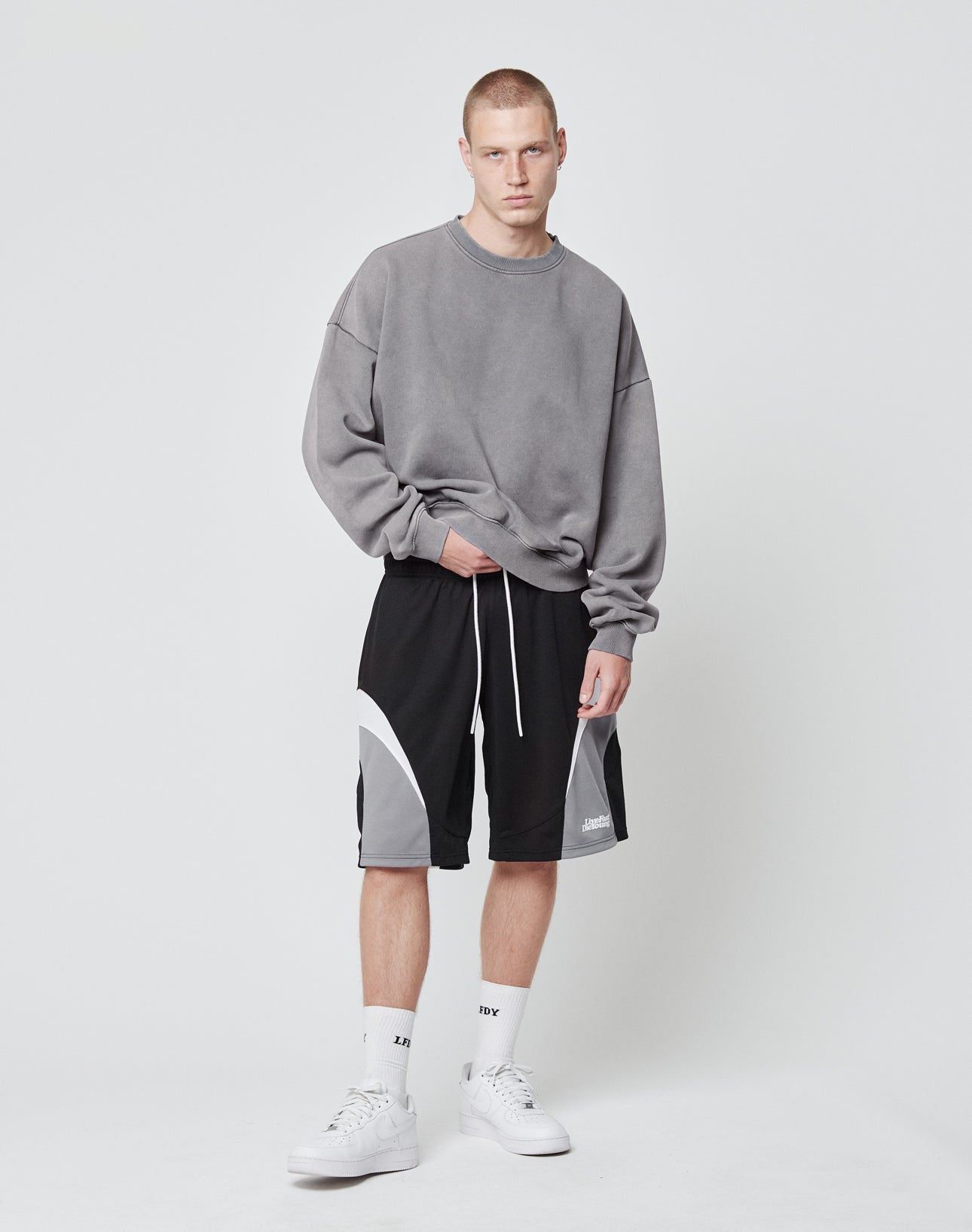 A young man with short hair and a neutral expression stands against a white background, wearing LIVE FAST DIE YOUNG Basic Trackshorts with an elastic waistband, a gray oversized sweatshirt, white socks, and white sneakers.