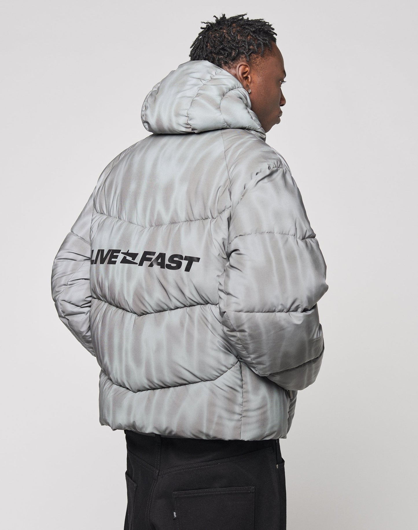 A person is seen from the back wearing the Live Fast Puffer Jacket by LIVE FAST DIE YOUNG in light gray with a water-repellent finish, branded hood piping, and LIVE FAST boldly printed across the upper back. The jacket has an oversized fit.