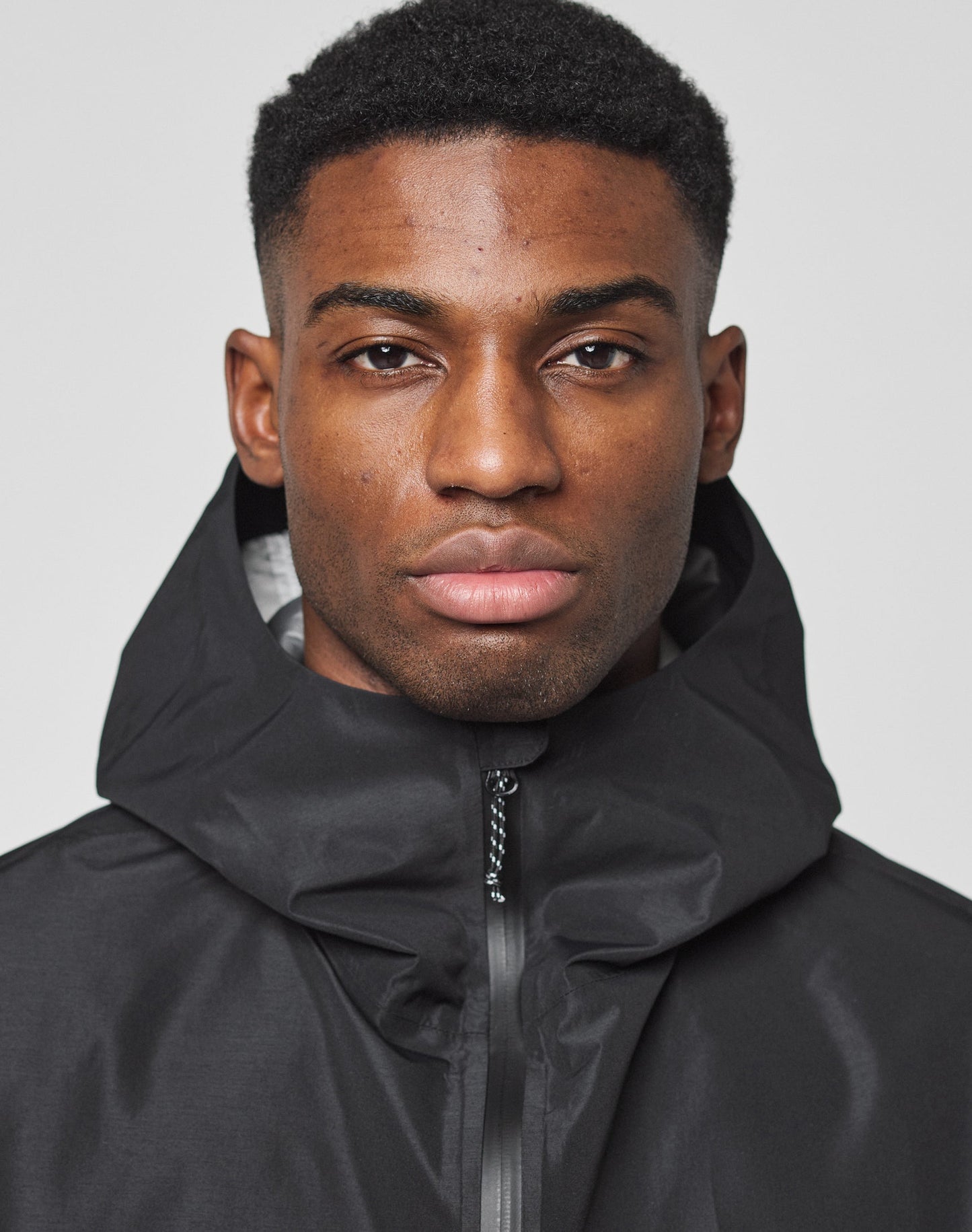 A man with short black hair wears the LIVE FAST DIE YOUNG Live Fast Performance Rain Jacket, featuring wasserdichte Nähte, zipped to the top, and looks neutrally at the camera against a plain light background.