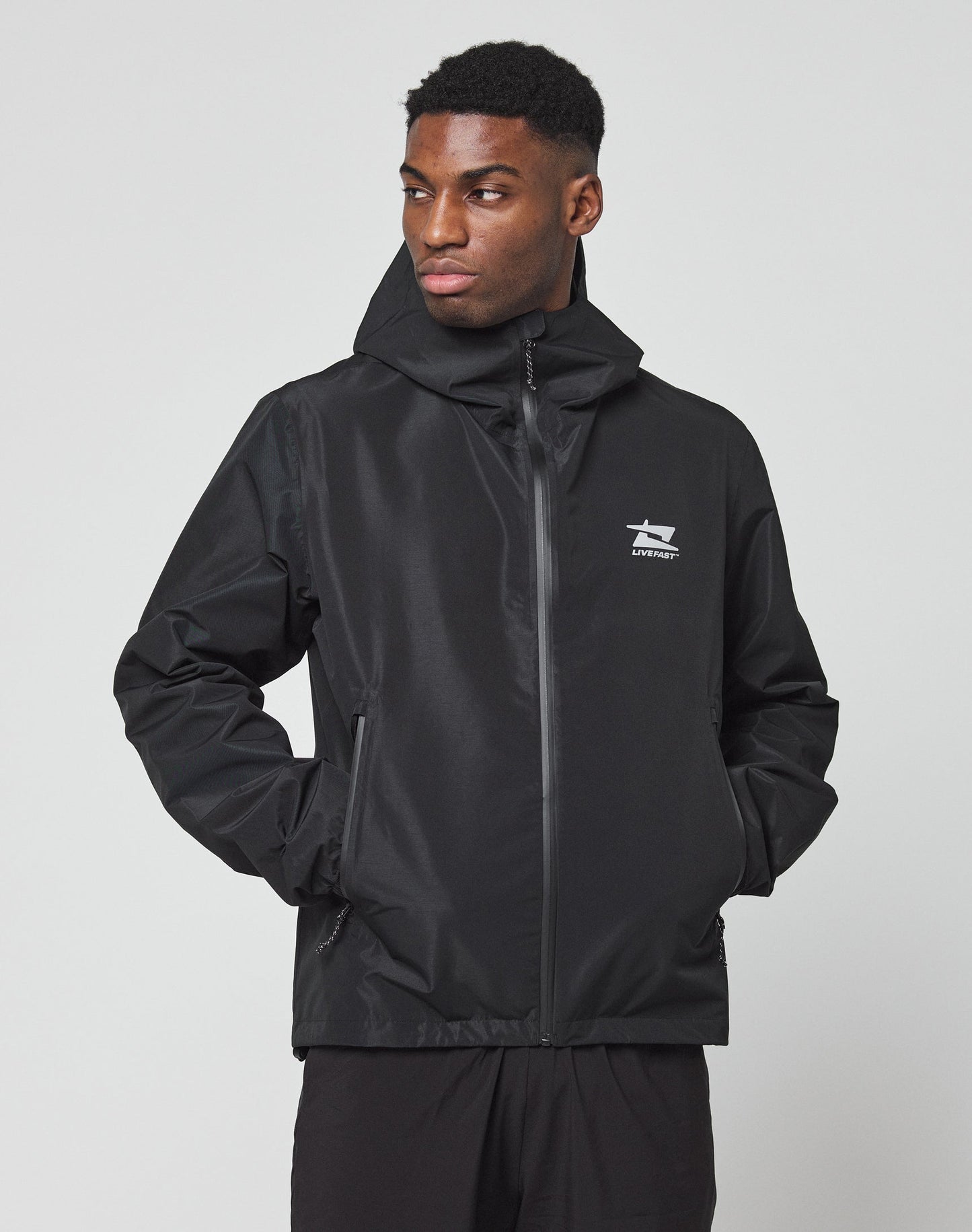 A man wears the Live Fast Performance Rain Jacket by LIVE FAST DIE YOUNG, featuring a black zip-up design, regular fit, elastic cuffs, and a white chest logo, standing with hands in pockets and looking left against a plain light background.