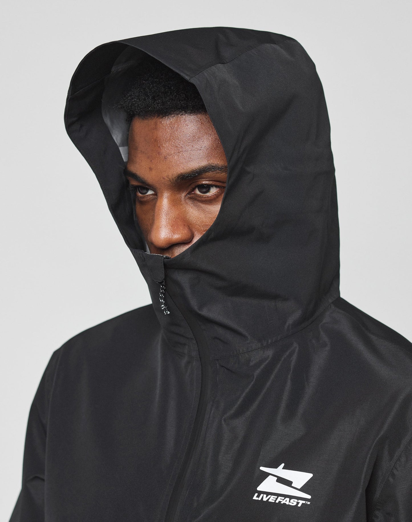 A person wears the LIVE FAST DIE YOUNG Live Fast Performance Rain Jacket in black with the hood up, partially covering their face. This regular fit jacket has a white LIVE FAST logo on the chest and elastic cuffs. The background is plain light gray.