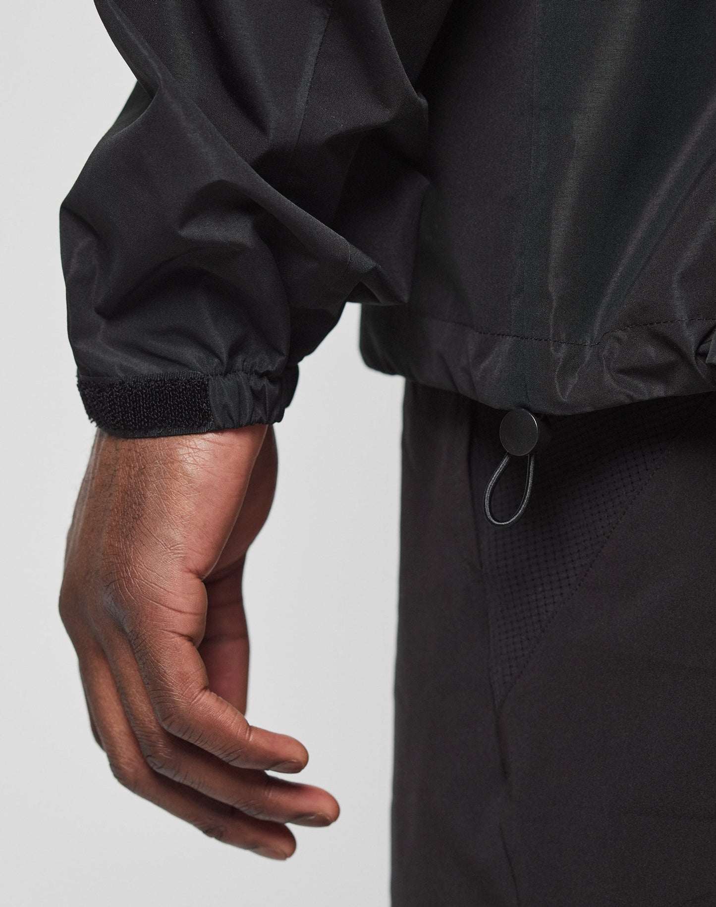 A close-up shows a person’s left hand and arm wearing the LIVE FAST DIE YOUNG Live Fast Performance Rain Jacket with elastic cuffs, an adjustable hem drawstring, regular fit, and black pants against a plain light background.