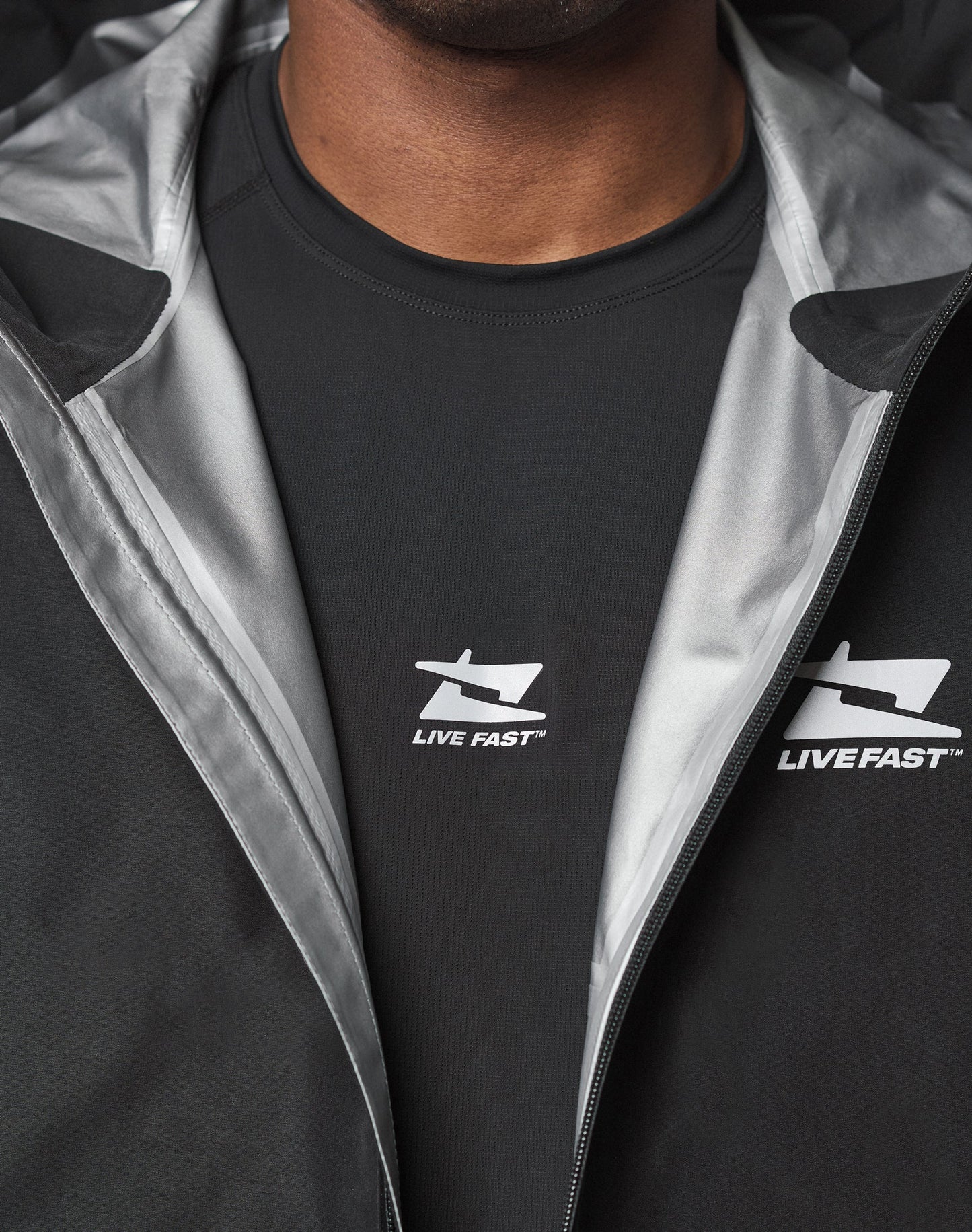 Close-up of a person wearing the black Live Fast Performance Rain Jacket by LIVE FAST DIE YOUNG, featuring Elastische Ärmelöffnung and partially unzipped to reveal a black shirt with the LIVE FAST logo and arrow design.
