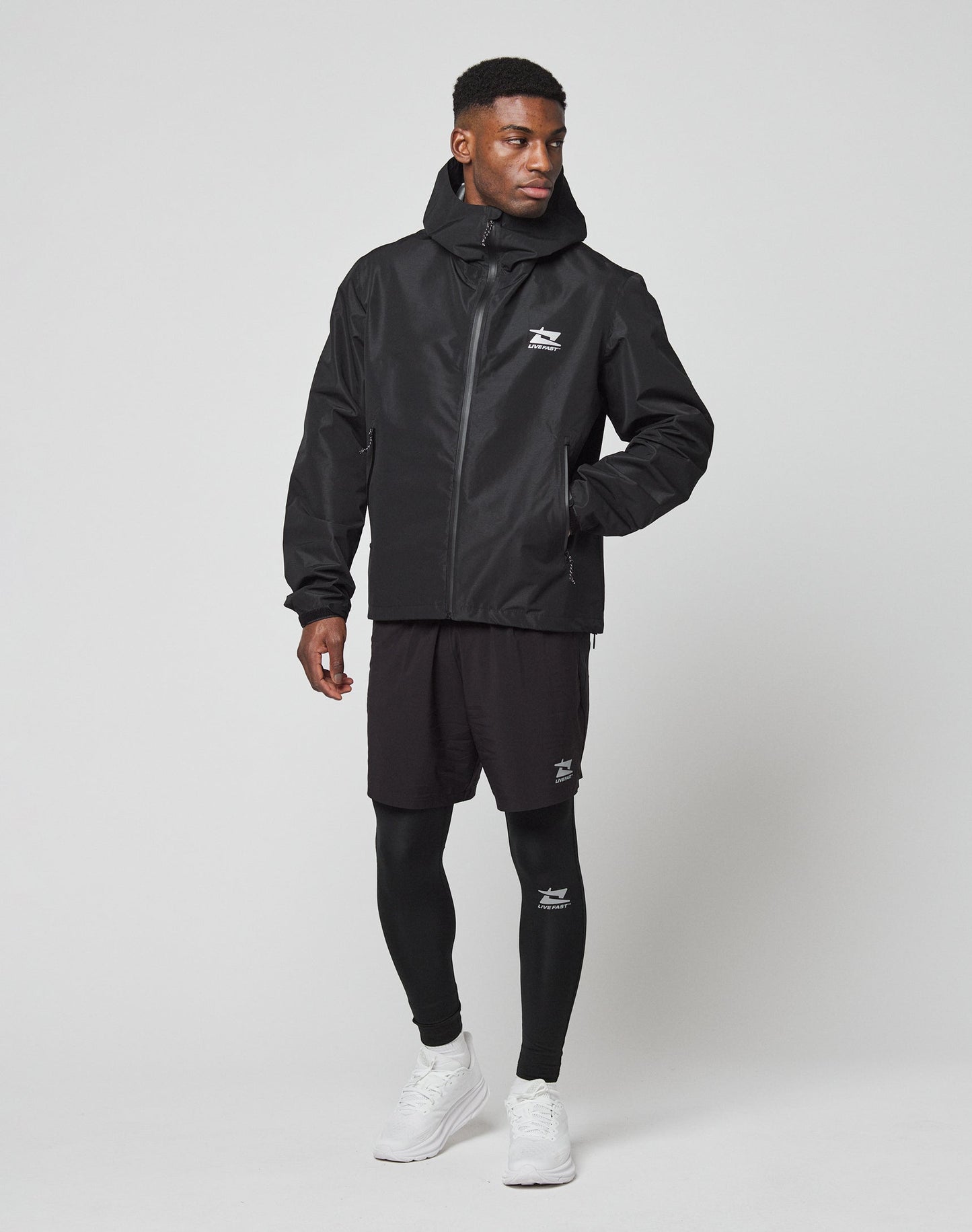 A man stands against a plain background wearing the LIVE FAST DIE YOUNG Live Fast Performance Rain Jacket with Wasserdichte Nähte, black shorts, black leggings, and white sneakers. He looks to the side with one hand in his pocket.