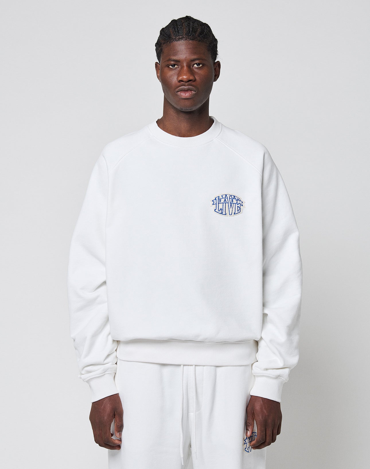 A person faces forward wearing a LIVE FAST DIE YOUNG LFDY Radiator Sweater in oversize white with matching pants. The Baumwolle Polyester sweater has a small blue LEARN TO LIVE graphic on the left chest. The background is plain light gray.