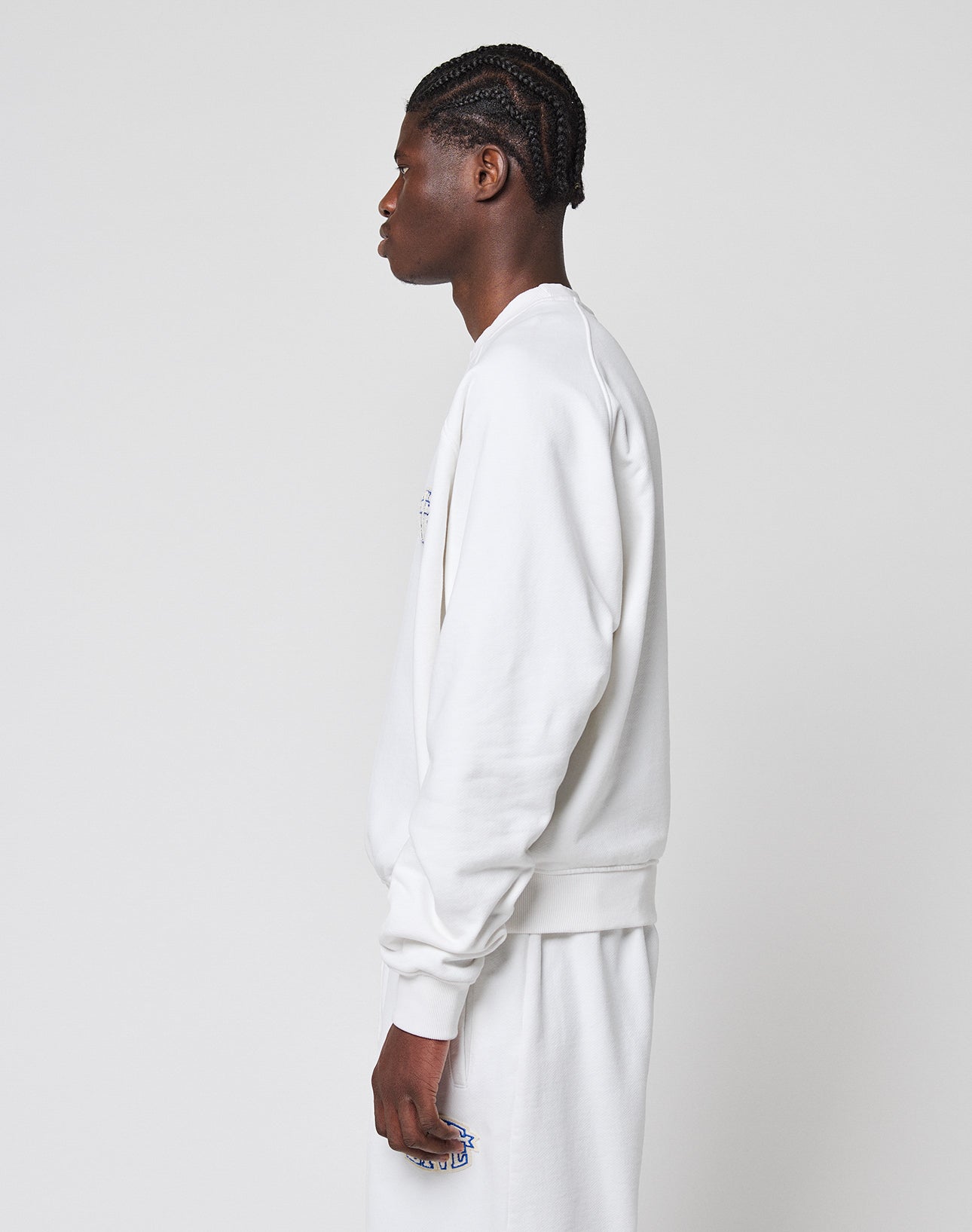 A person with braided hair stands in profile against a plain background, wearing an oversize fit white LFDY Radiator Sweater and matching white pants from LIVE FAST DIE YOUNG.