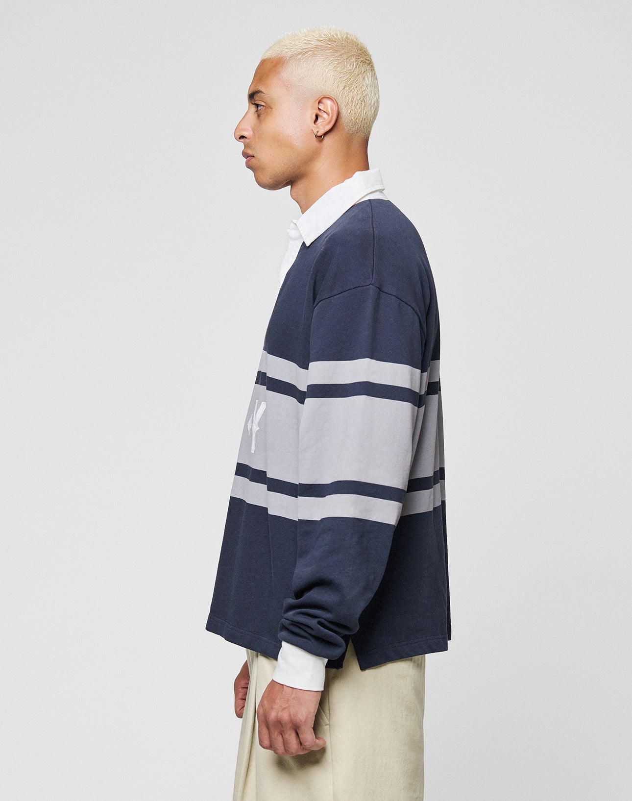 A person with short, bleached blonde hair stands in profile against a plain background, wearing the LFDY Striped Polo by LIVE FAST DIE YOUNG over a white collared shirt and beige cotton twill pants.