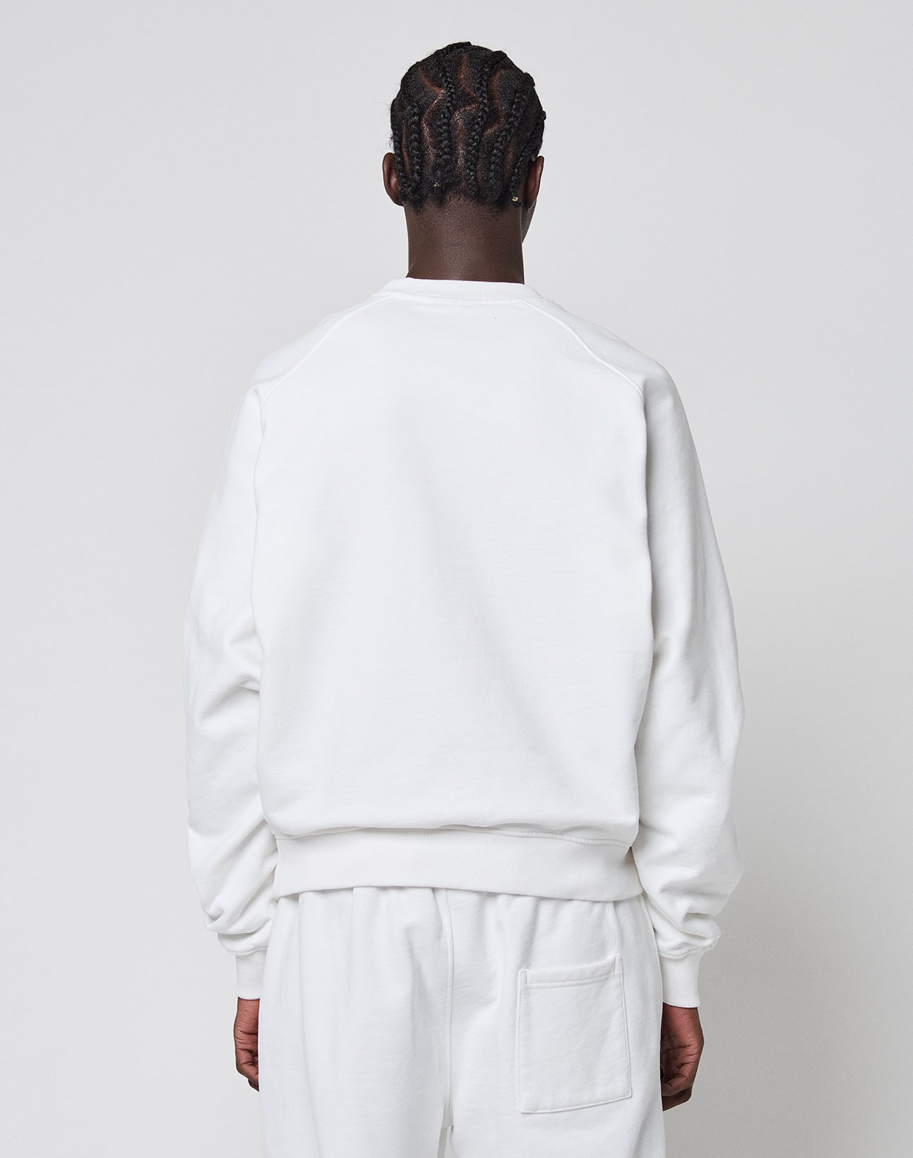 A person with braided hair, facing away from the camera, wears an oversize white LFDY Radiator Sweater and matching pants by LIVE FAST DIE YOUNG, both in a soft Baumwolle Polyester blend, set against a plain light gray background.