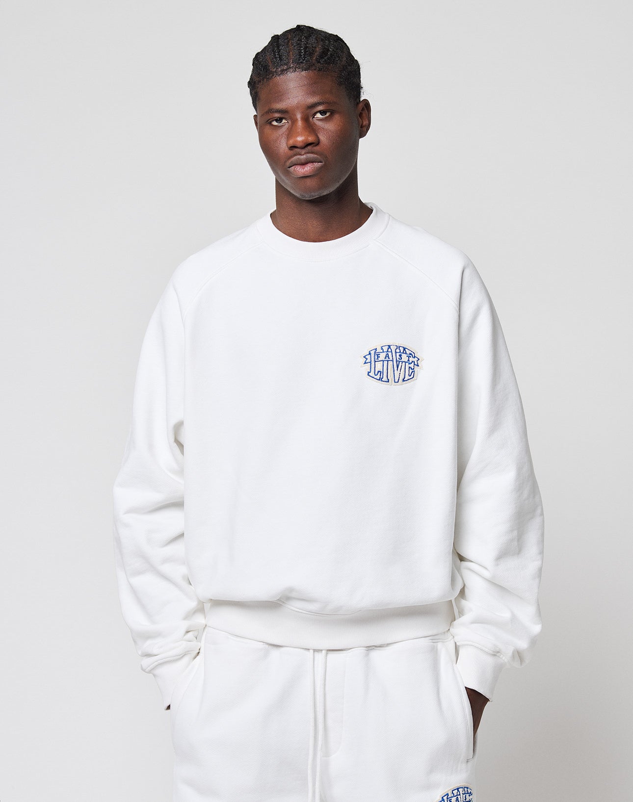 A person models the white LFDY Radiator Sweater by LIVE FAST DIE YOUNG, styled with matching sweatpants, against a light gray background. The sweater features an oversize fit and a small blue chenille patch logo on the chest.
