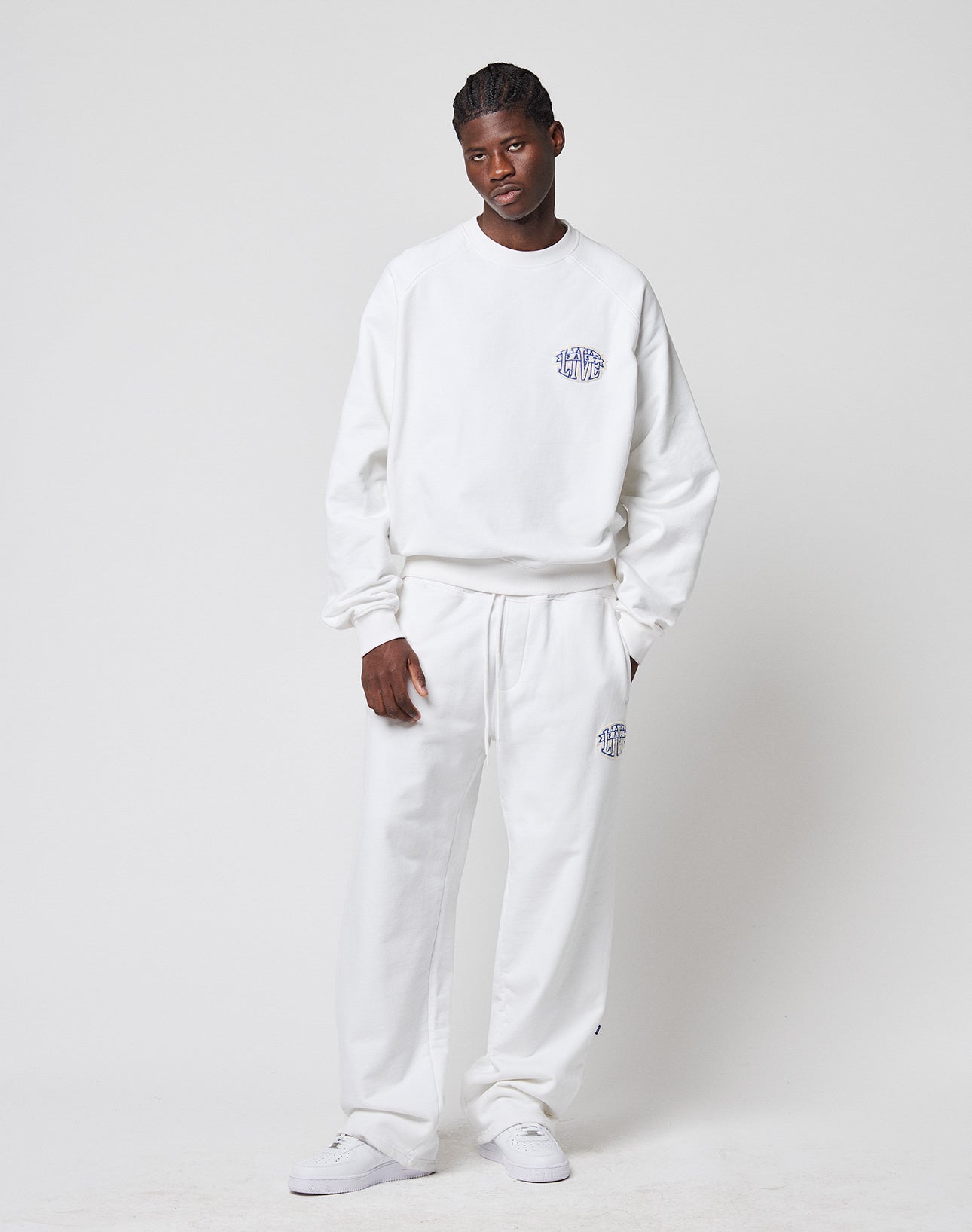 A person with short braided hair and a relaxed posture models LIVE FAST DIE YOUNG’s LFDY Radiator Sweatpants and matching oversized white sweatshirt, featuring small blue and purple logos, chenille patches, elastic waists, and white sneakers.