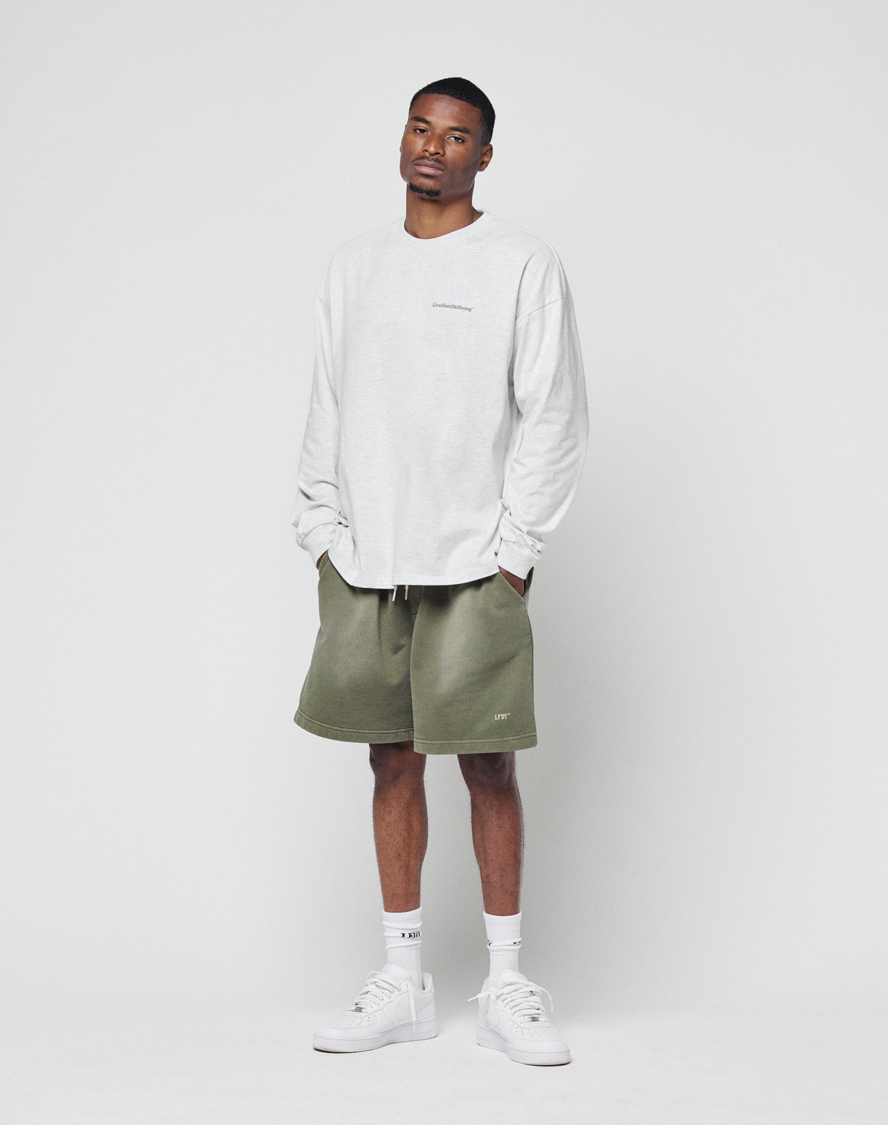 Against a plain white backdrop, a man wears the LIVE FAST DIE YOUNG Basic Longsleeve in light gray with an oversize fit, styled with olive green shorts, white crew socks, and white sneakers. His hands are in his pockets and he has a neutral expression.