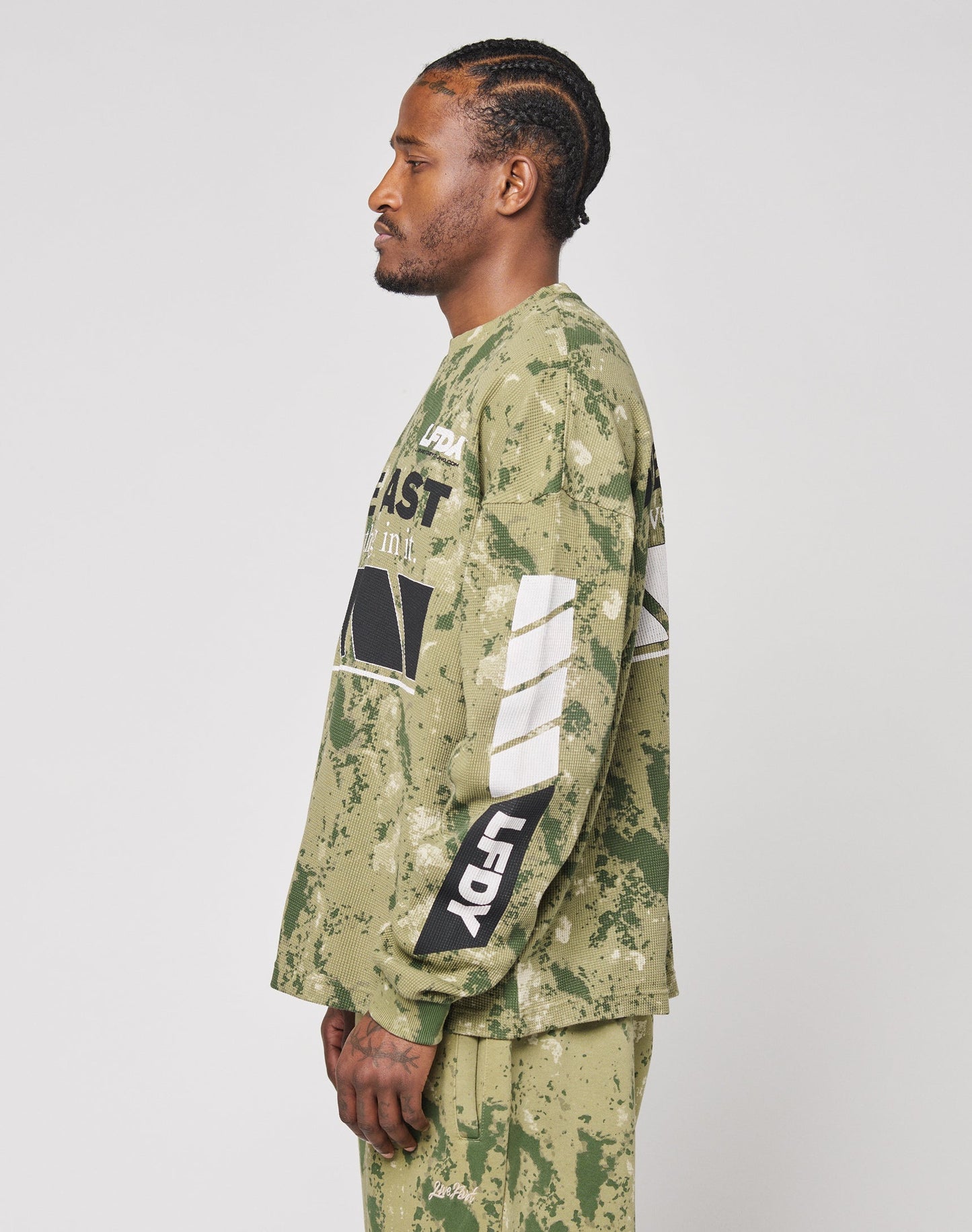 A man with cornrow braids wears the LIVE FAST DIE YOUNG LFDY Racing Waffle Longsleeve—an oversize 100% Baumwolle shirt with bold black and white front graphics—paired with matching pants, standing in profile against a light background.