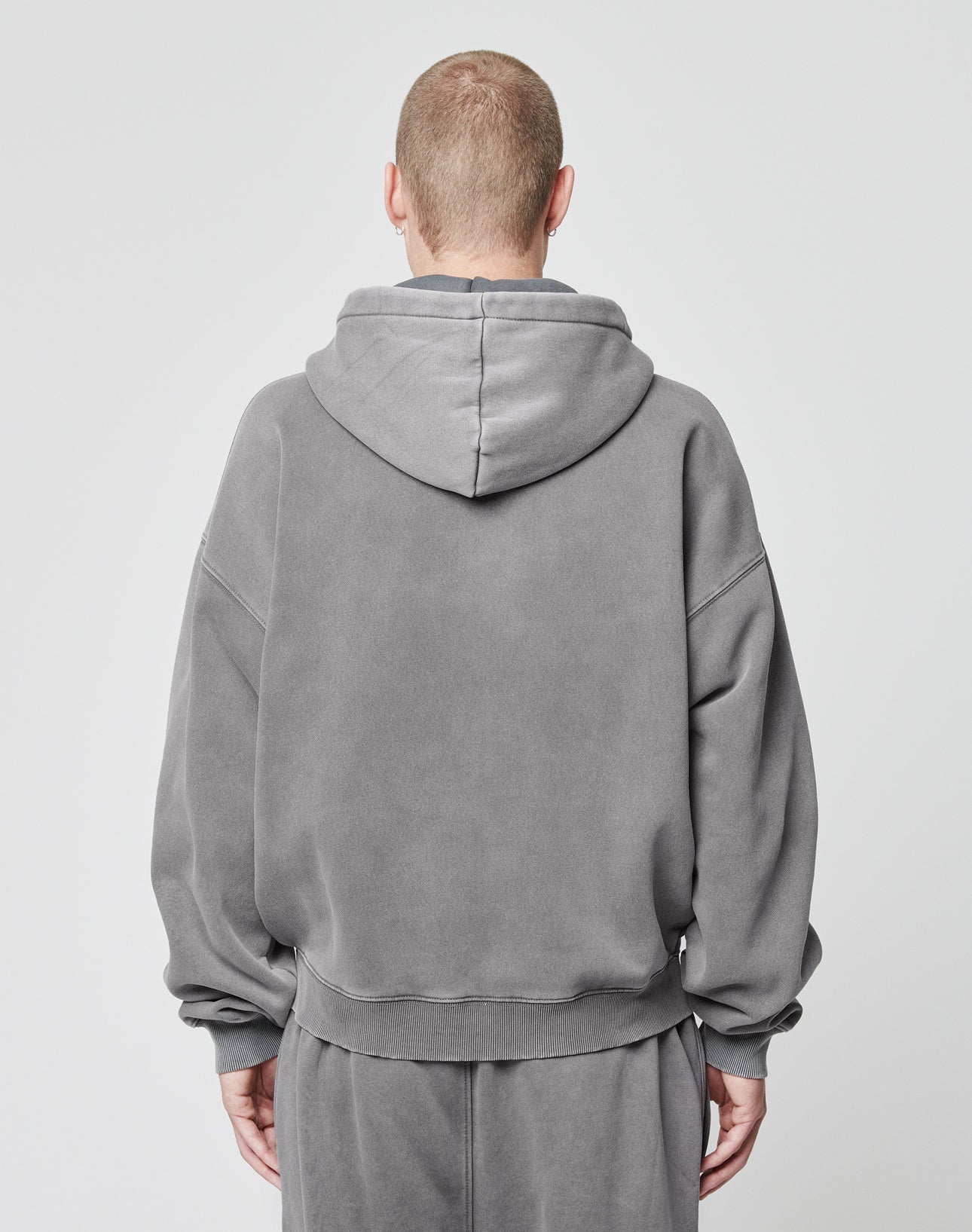 A person with short hair faces away from the camera, wearing a loose-fitting LIVE FAST DIE YOUNG Basic Script Hooded Zip and matching sweatpants in vintage wash against a plain light background.