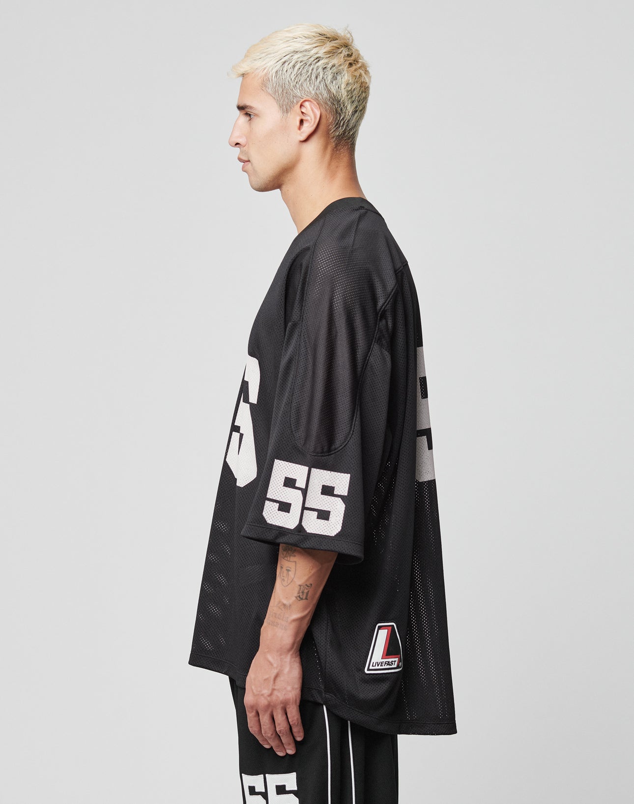 A person with short, bleached blonde hair stands in profile wearing an oversize fit black LIVE FAST DIE YOUNG LFDY Mesh Jersey with the number 55 on the sleeve and shorts against a plain light gray background.