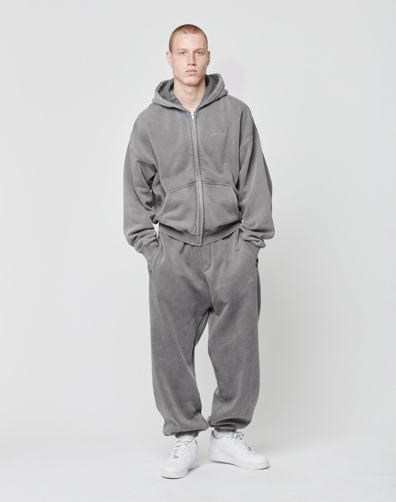 Vor neutralem Hintergrund trägt eine Person das oversized Basic Script Hooded Zip von LIVE FAST DIE YOUNG im Vintage-Look zu grauen Sweatpants und weißen Sneakern. Sie hat kurze Haare, neutrale Miene und die Hände in den Taschen.